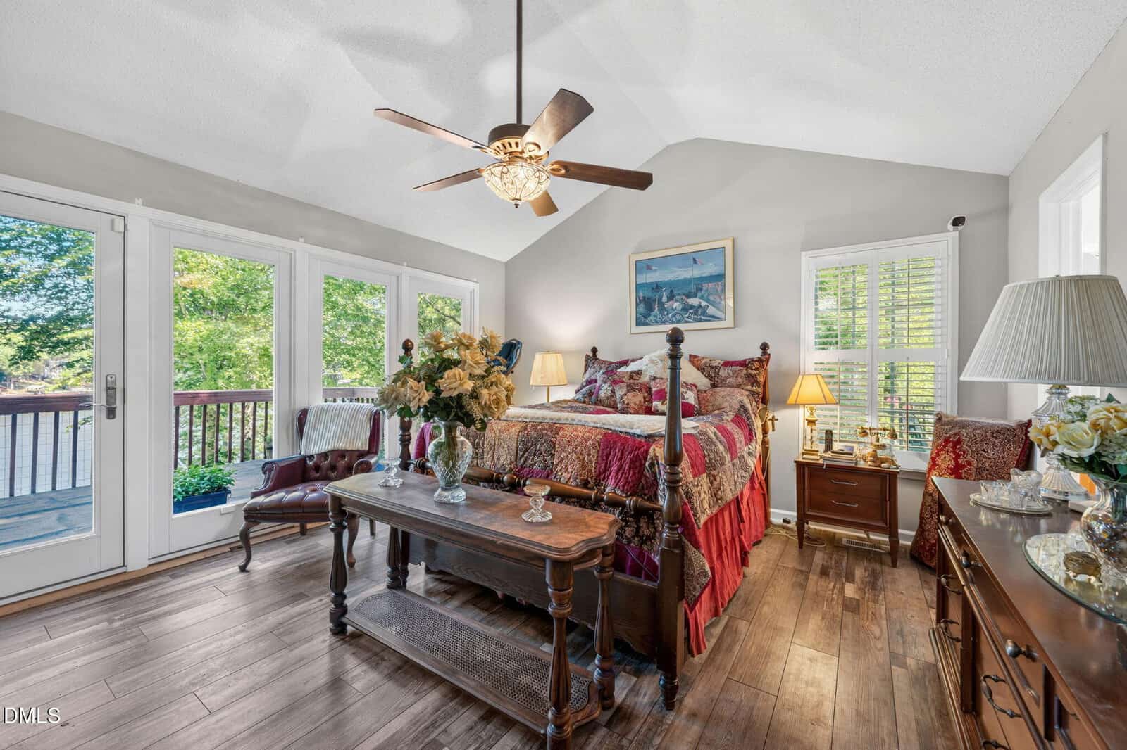 A cozy bedroom with a wooden floor, four-poster bed with colorful bedding, a ceiling fan, vintage furniture, lamps, flowers, and large windows opening to a deck with greenery outside.