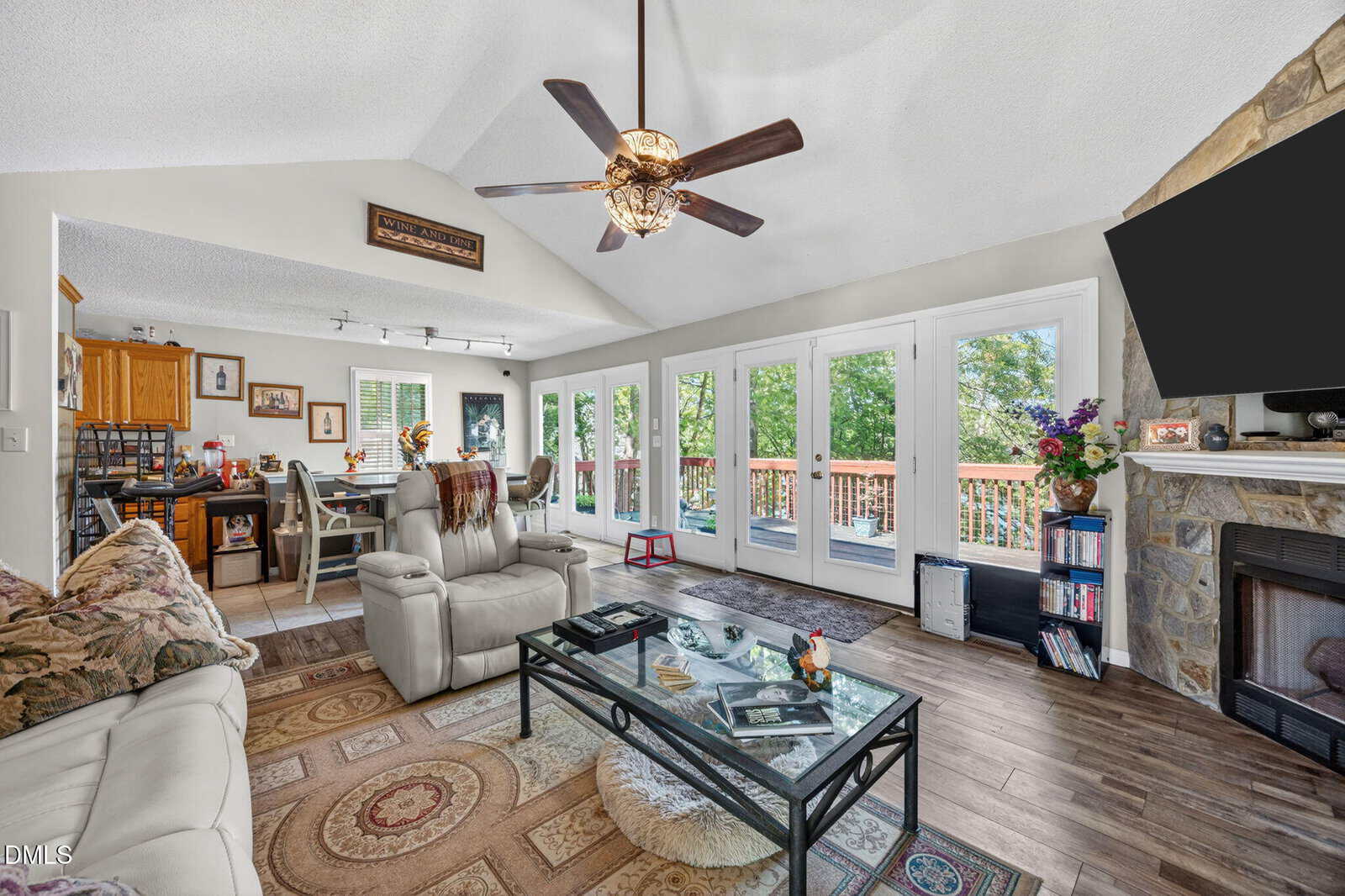 Bright living room with beige sofas, a glass coffee table, and a ceiling fan. Large windows and glass doors open to a deck with trees outside. A stone fireplace and TV are in one corner, and a kitchen is in the background.