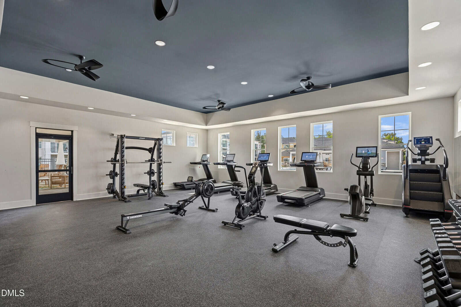 Modern gym room with various exercise equipment including treadmills, rowing machines, elliptical machines, weight racks, and a squat rack. Large windows let in natural light, and the space has a dark ceiling and light walls.
