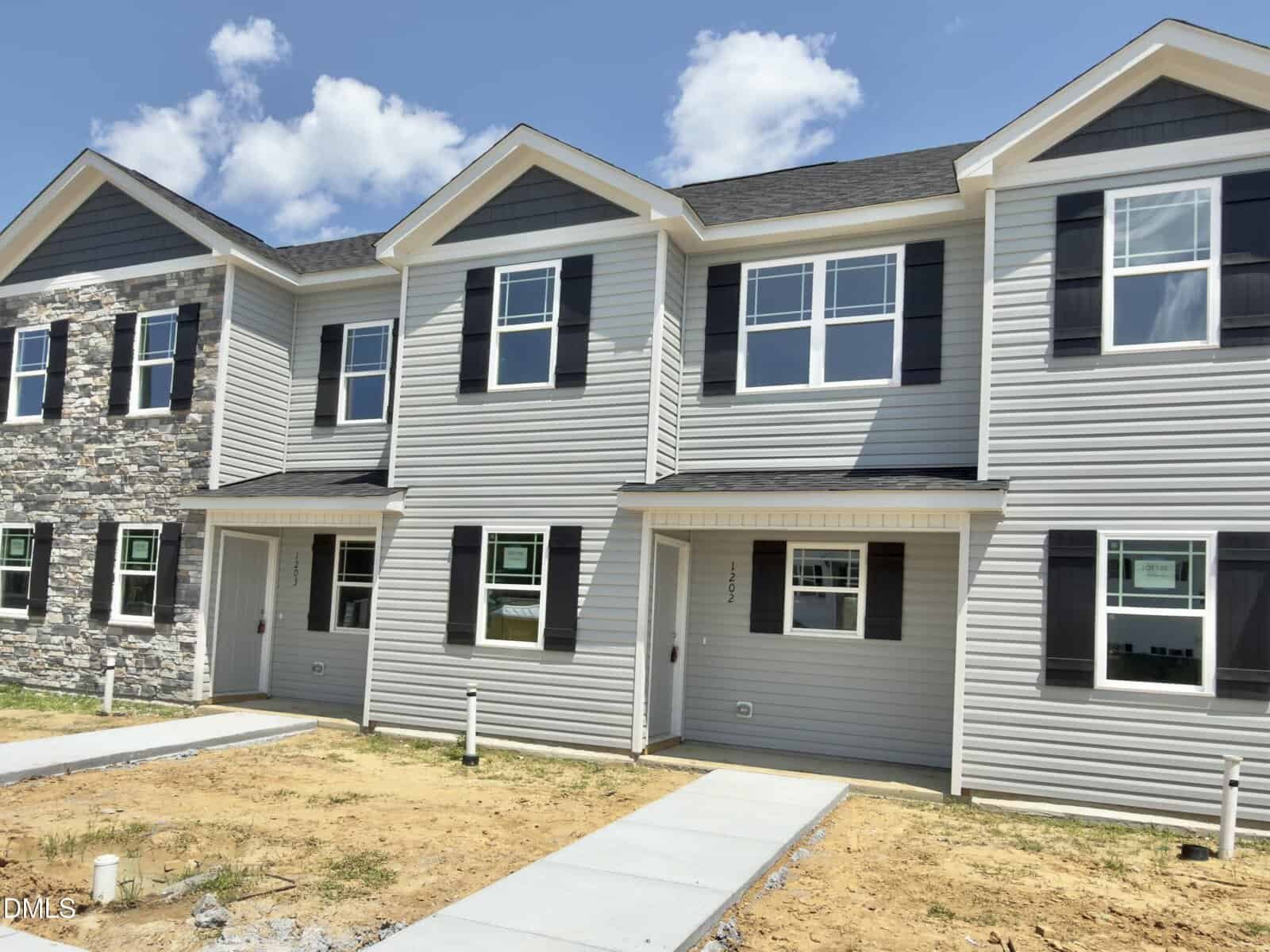 A row of newly constructed two-story townhouses on 1202 Brookrun Drive in Oxford features gray siding, black shutters, and white trim. The front yards are dirt and unfinished, with concrete walkways leading to each entrance. The sky is partly cloudy.