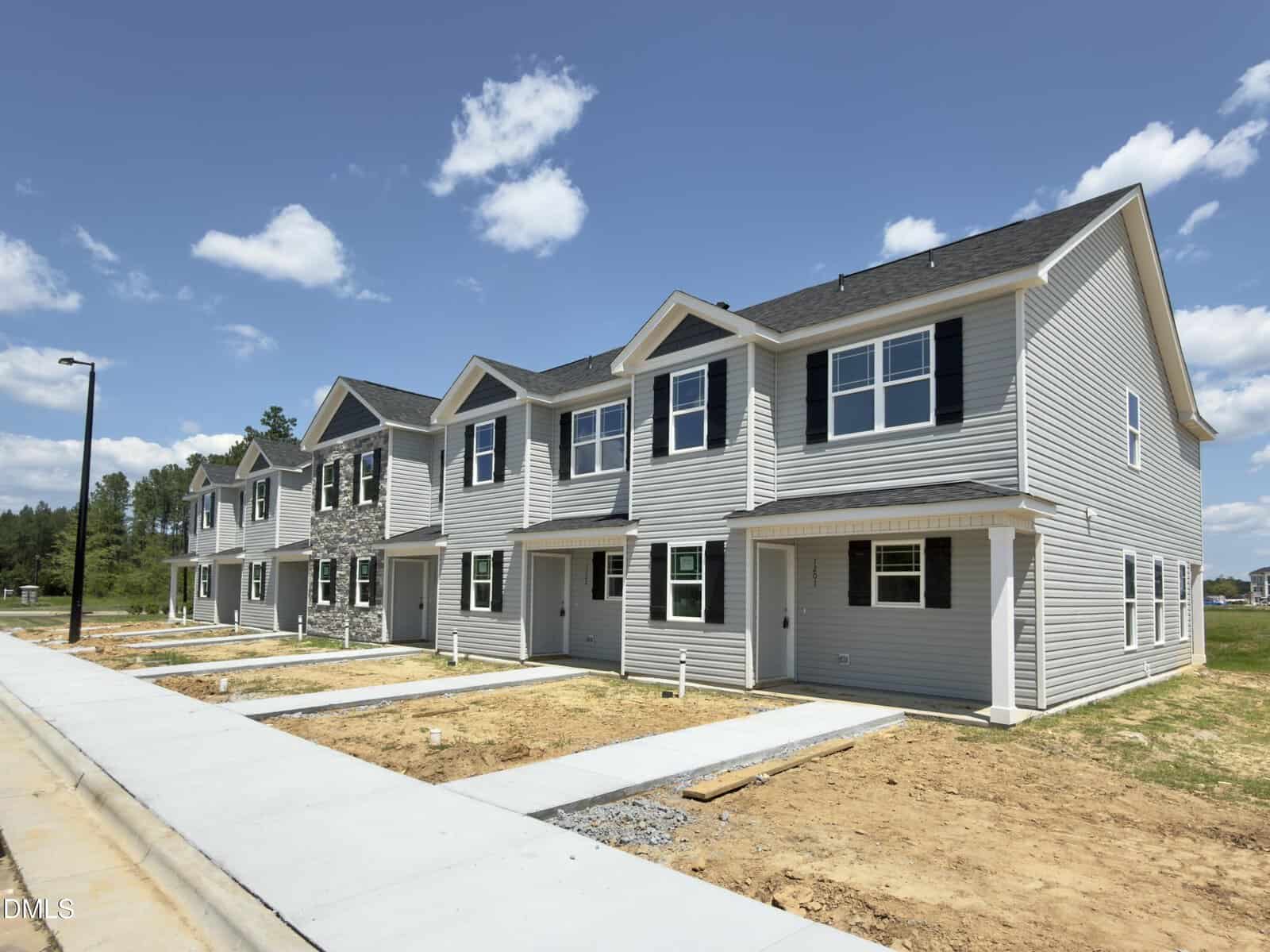 A row of newly constructed two-story townhouses with gray siding, white trim, and black shutters. The houses have concrete driveways and unfinished yards. Blue sky with scattered clouds is visible above.