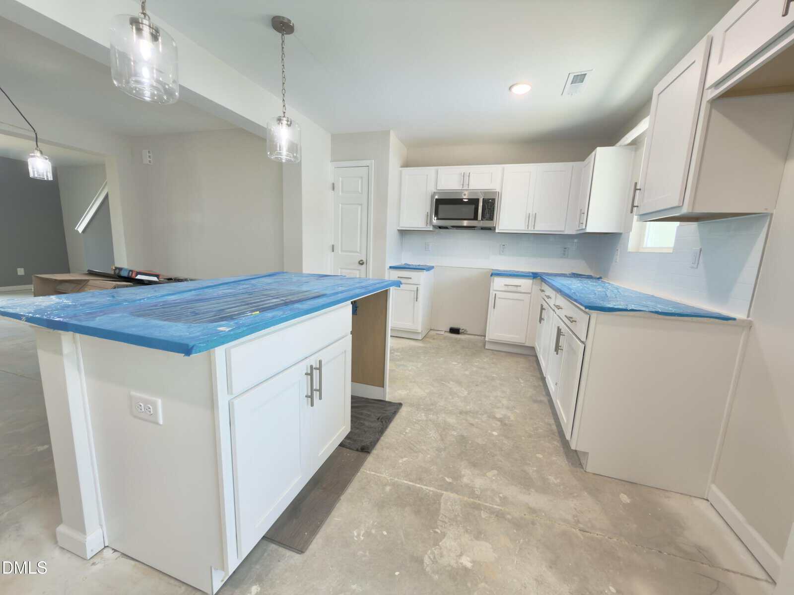 A modern kitchen with white cabinets, blue protective cover on countertops, stainless steel appliances, and unfinished floors. Two pendant lights hang above the island. The space appears to be under renovation or construction.