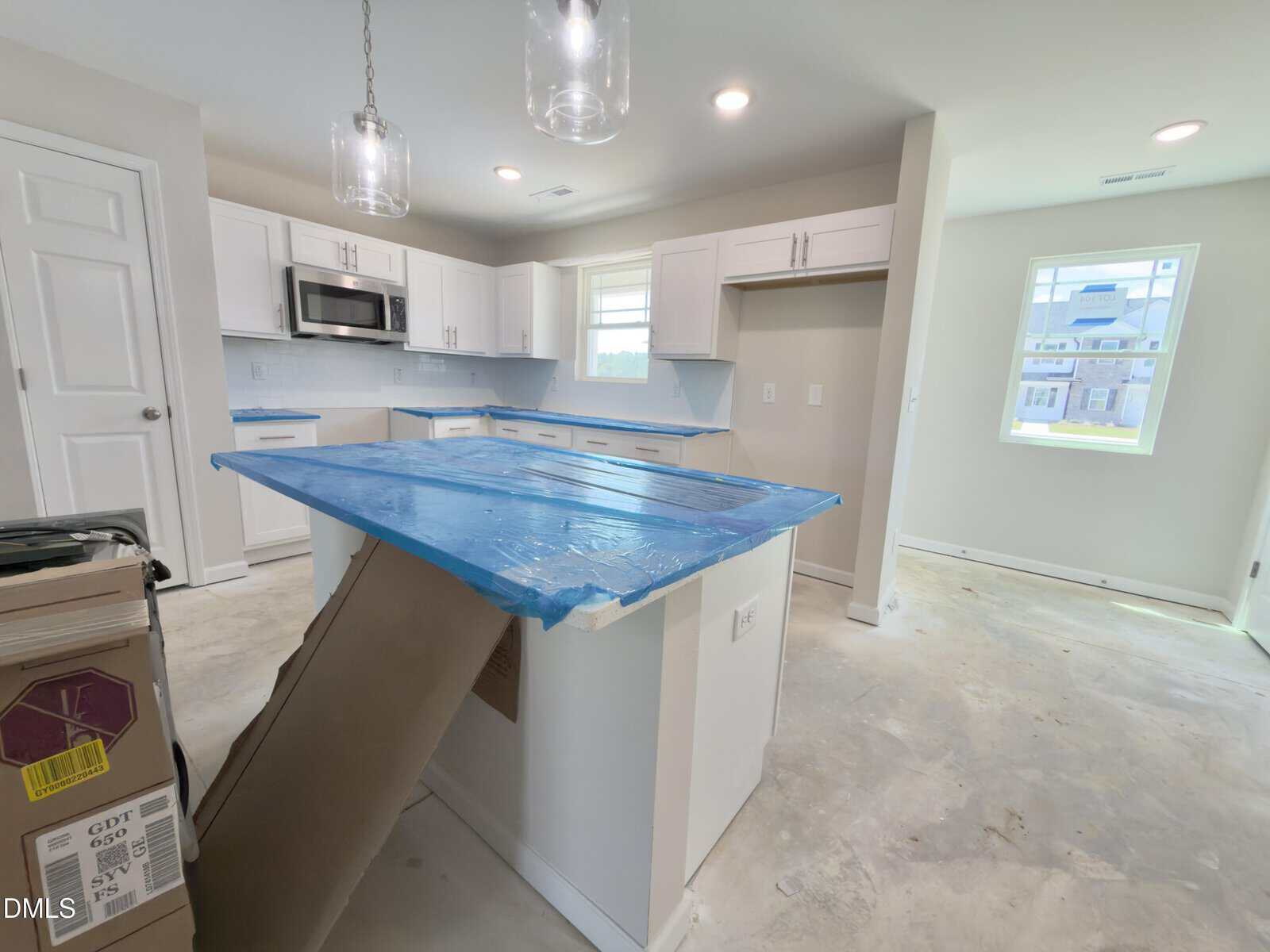 A kitchen under construction with white cabinets, a blue plastic-covered island, unfinished floors, and boxes on the left. A microwave is built into the cabinets, and light fixtures hang from the ceiling. Natural light enters through two windows.