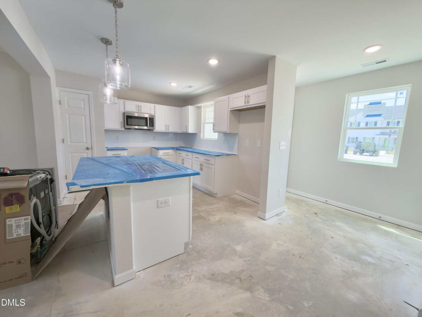 Unfinished kitchen in a new home with white cabinets, blue protective film on counters, exposed concrete floor, a kitchen island, and a window letting in daylight. Appliances appear uninstalled.