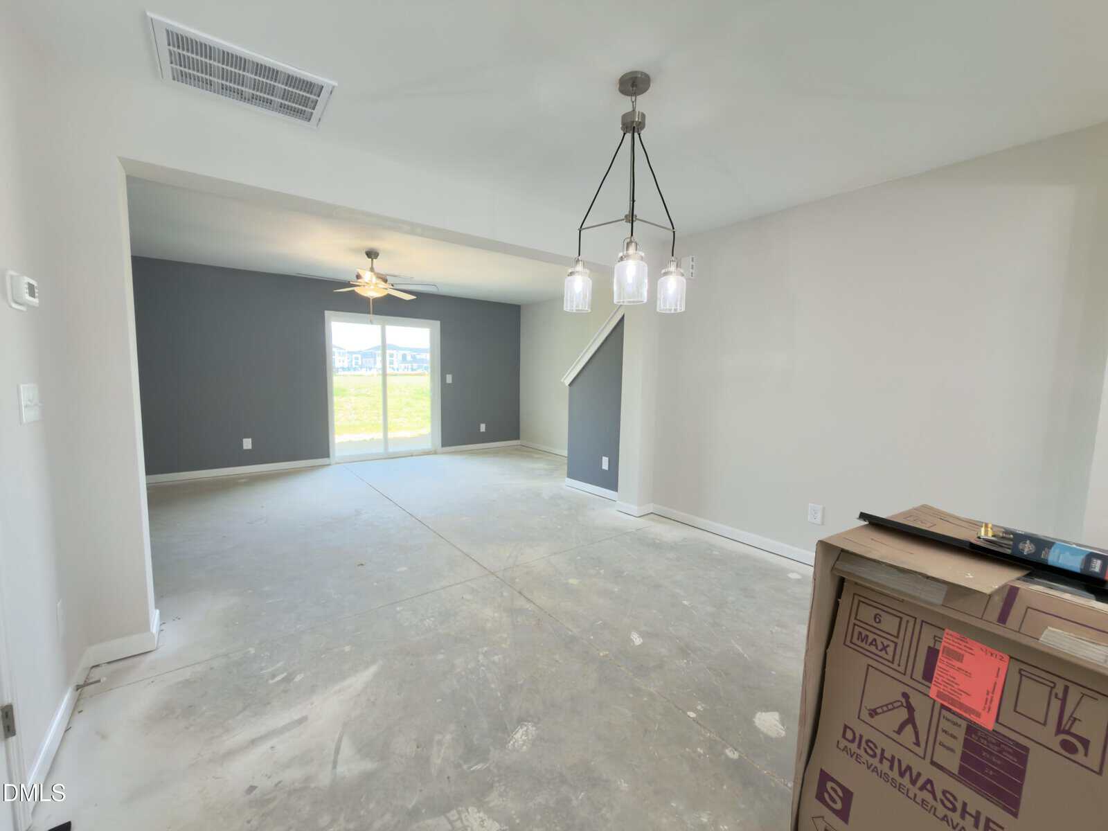 An empty, unfurnished room with gray and white walls, unfinished concrete floor, a ceiling fan, modern light fixture, and a cardboard box labeled “dishwasher” near the entrance. Sunlight enters through windows at the back.