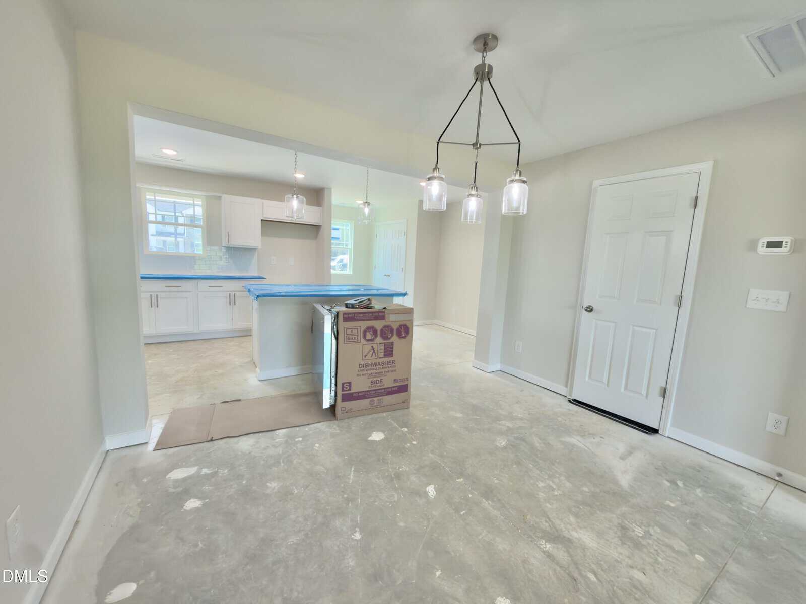 A room with unfinished floors, a box in the center, and a view into a kitchen with white cabinets. Three pendant lights hang above, and there is a closed white door and a wall thermostat on the right.