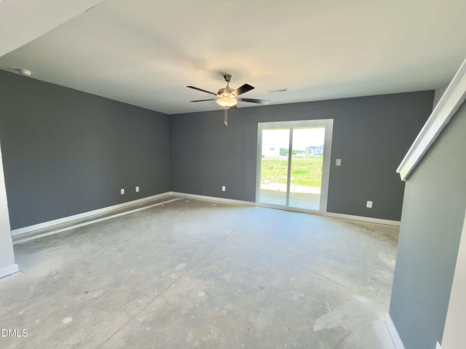 Empty room with gray walls, white trim, and concrete floor. A ceiling fan with lights is centered on the ceiling. Sunlight comes through glass sliding doors that open to a grassy outdoor area. Stairs are partially visible on the right.