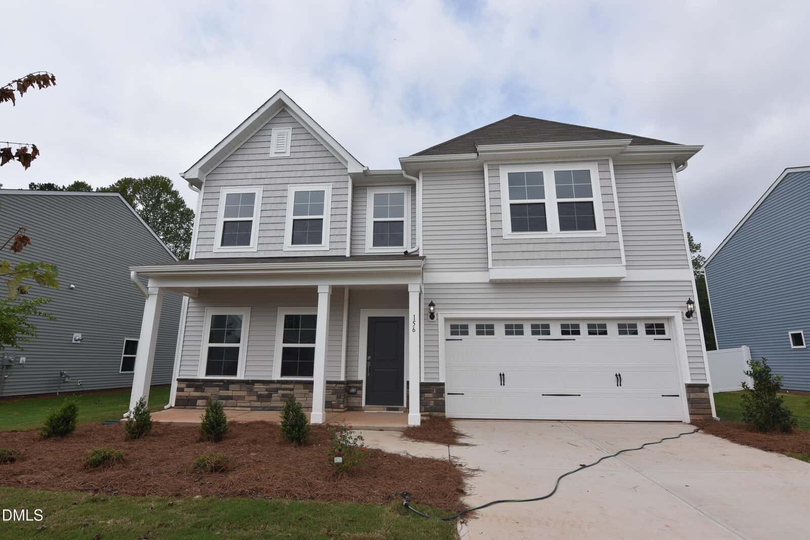 Two-story modern house with gray siding, white trim, and stone accents located at 156 Sugarmaple Way in Youngsville. There’s a covered front porch, double garage, and a driveway with a garden hose on the newly landscaped front yard under a partly cloudy sky.