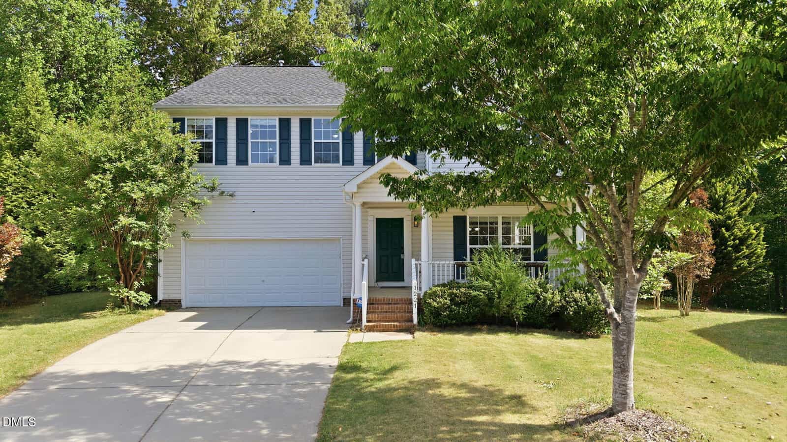 A two-story suburban house with white siding, dark shutters, a covered front porch, and an attached two-car garage. A large tree and neatly trimmed shrubs are in the front yard, with a driveway leading to the garage.