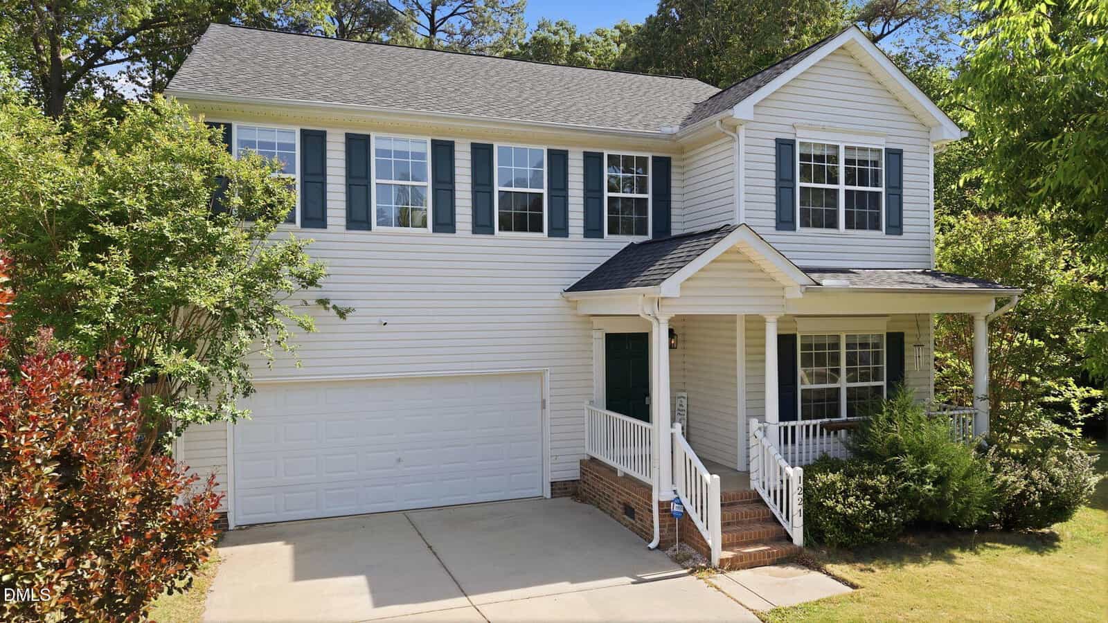 Two-story white house with dark shutters, a covered front porch, and an attached two-car garage. The home has a brick front step, surrounded by green trees and shrubs, with a concrete driveway.
