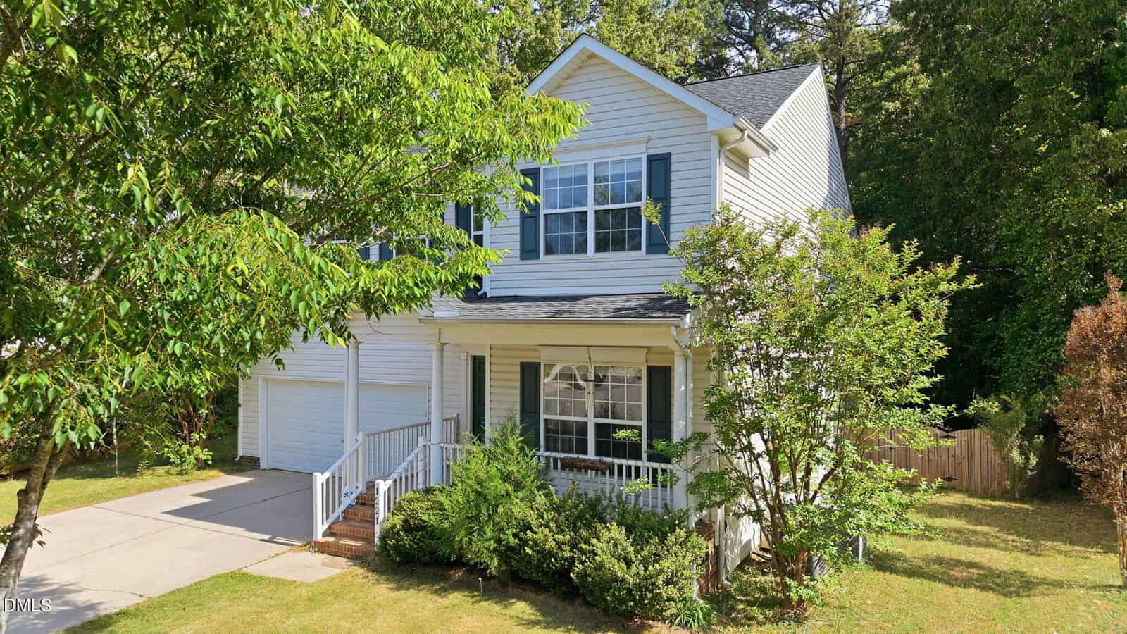 Two-story white house with green shutters, a covered front porch, attached two-car garage, and a well-maintained front yard with trees and shrubs. A driveway leads to the garage.
