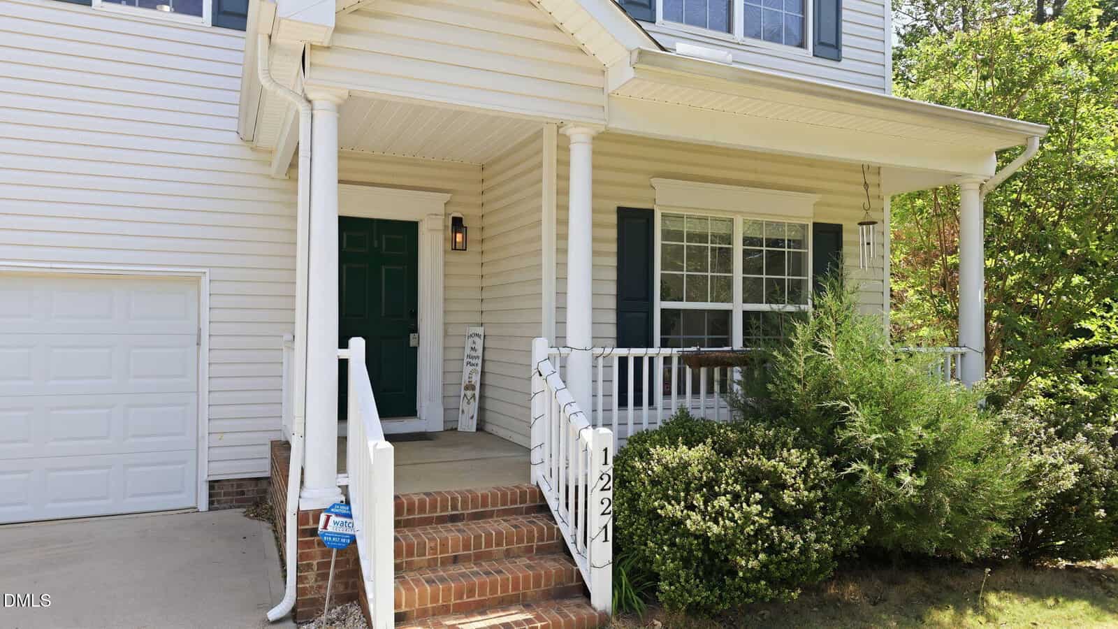 A front porch of a white house with green shutters, a dark green door, white columns, brick steps, and shrubs along the edge. The house number 1221 is visible near the steps.