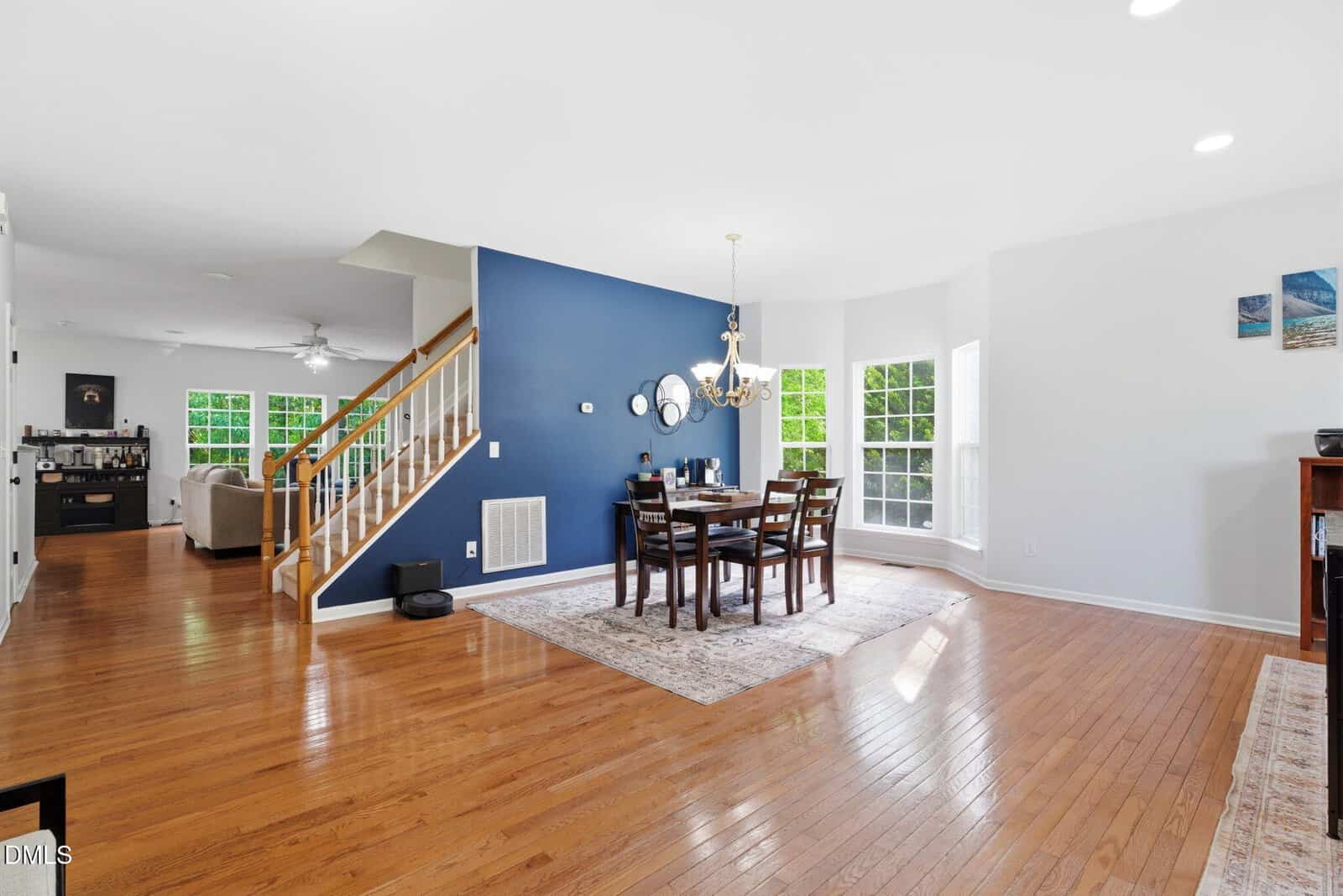 Open dining and living area with hardwood floors, a blue accent wall, modern chandelier, wooden dining table, large windows letting in natural light, and a staircase leading upstairs. Living room and kitchen visible in the background.