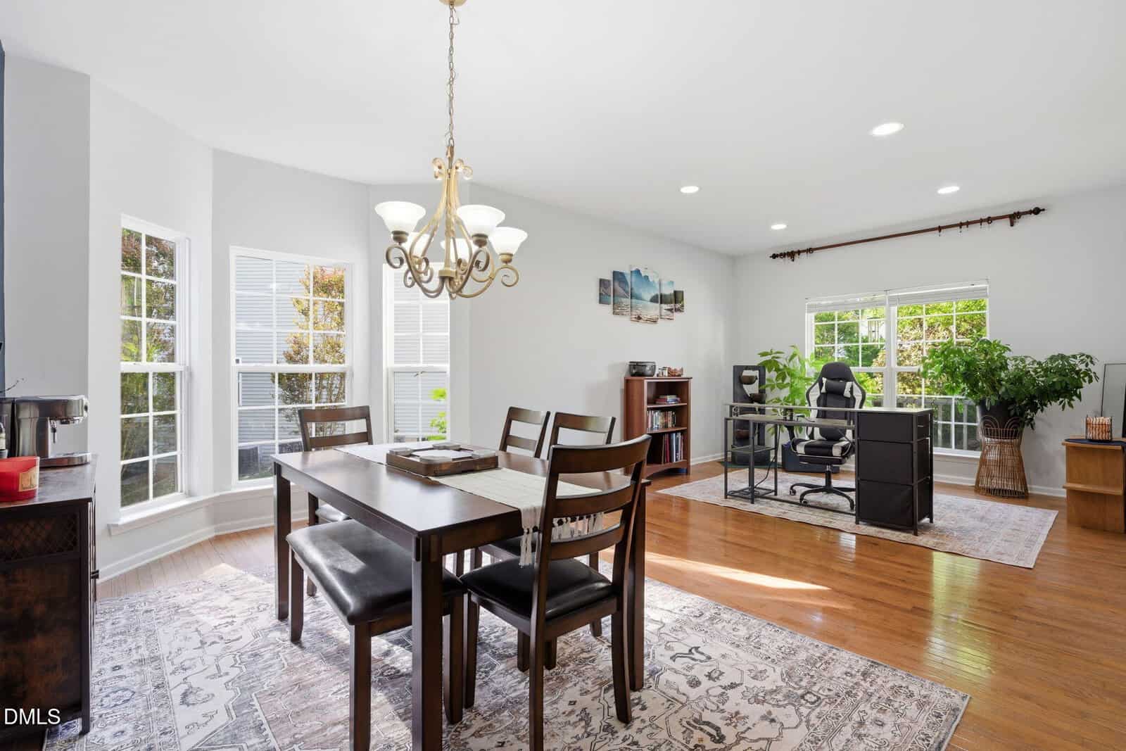 Bright dining and office area with large windows, wooden floor, chandelier above dining table, bookshelves, and desk with chair. Green plants add a touch of nature, and sunlight fills the modern, open space.