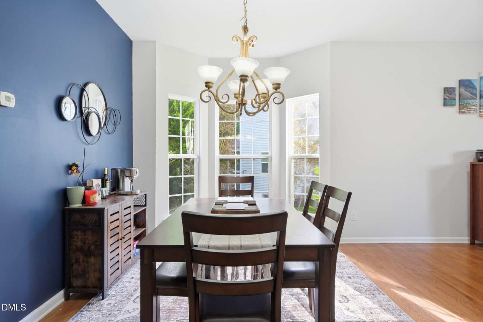 Bright dining room with a dark wood table, four chairs, a blue accent wall with mirrors, a chandelier, and tall windows letting in natural light. A sideboard with decor and a coffee station sits against the blue wall.