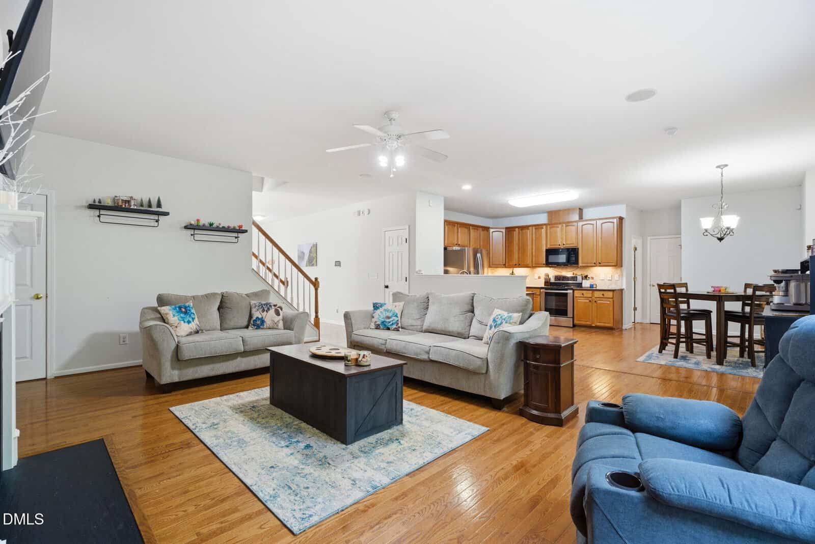 Open living room with two beige sofas, blue recliner, coffee table, and rugs; adjacent to a kitchen with wooden cabinets and a dining area with a dark table set on a hardwood floor. Stairs lead up near the kitchen.