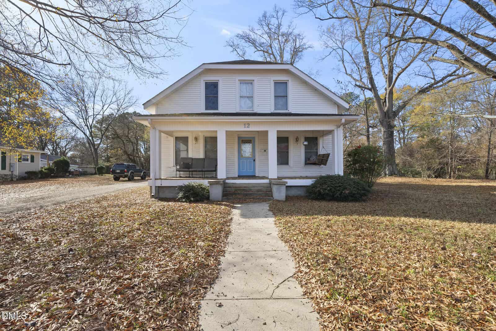 White two-story house with a blue front door, a covered porch, and two swings. Bare trees and autumn leaves cover the ground, and a curved walkway leads to the entrance. The sky is clear and blue.