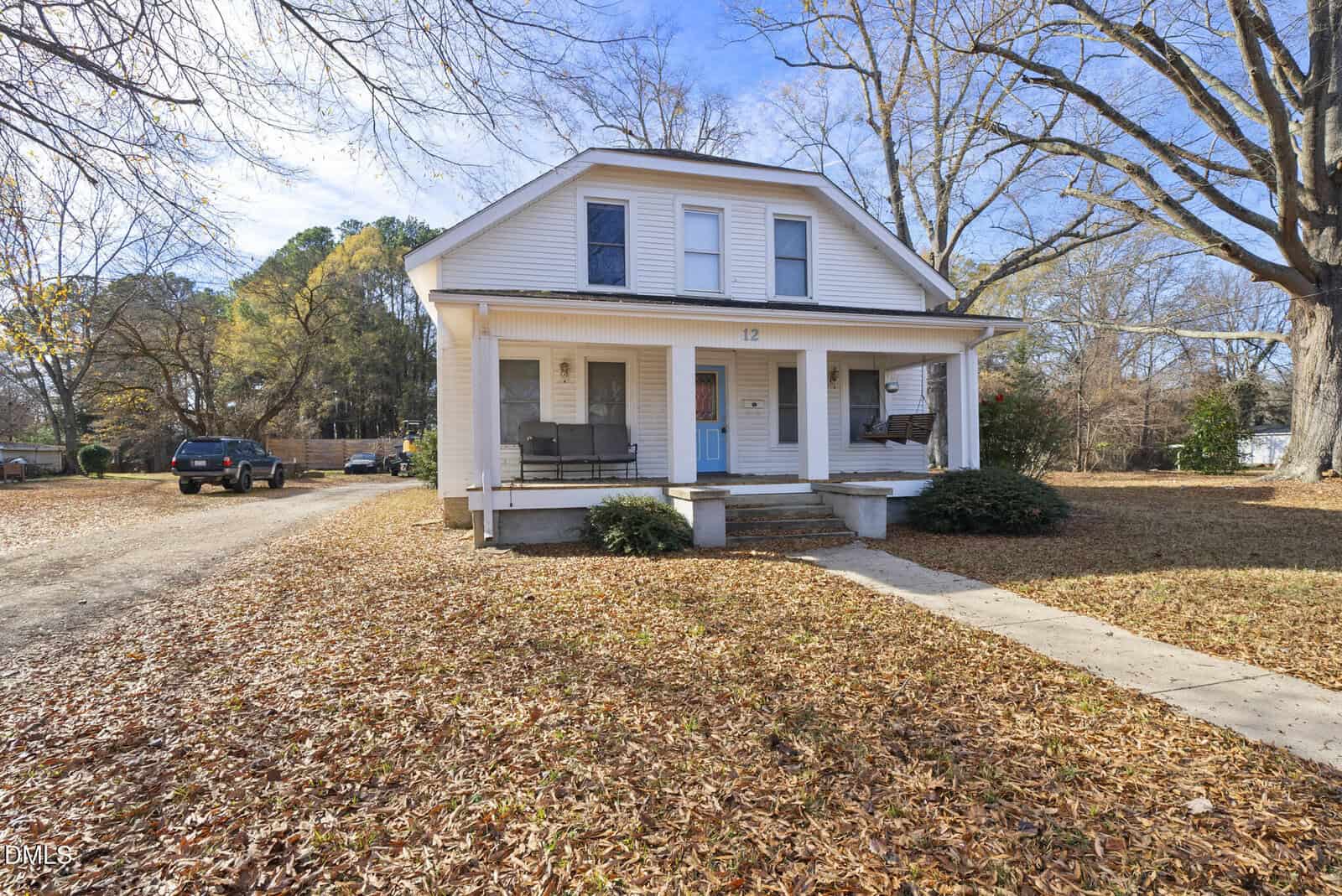 A white two-story house with a blue front door, two covered porches with swings, and a leaf-covered yard. A car is parked on a gravel driveway to the left, and large trees surround the property on a clear day.