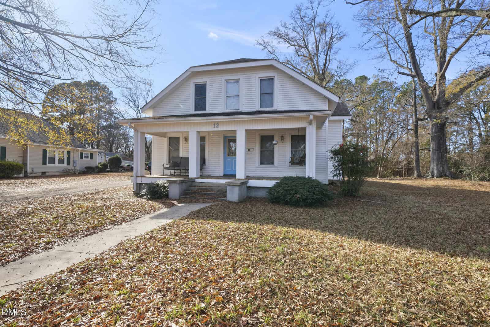 A white two-story house with a large front porch, light blue front door, and two pillars. The front yard is covered with autumn leaves, and tall trees surround the property under a clear sky.