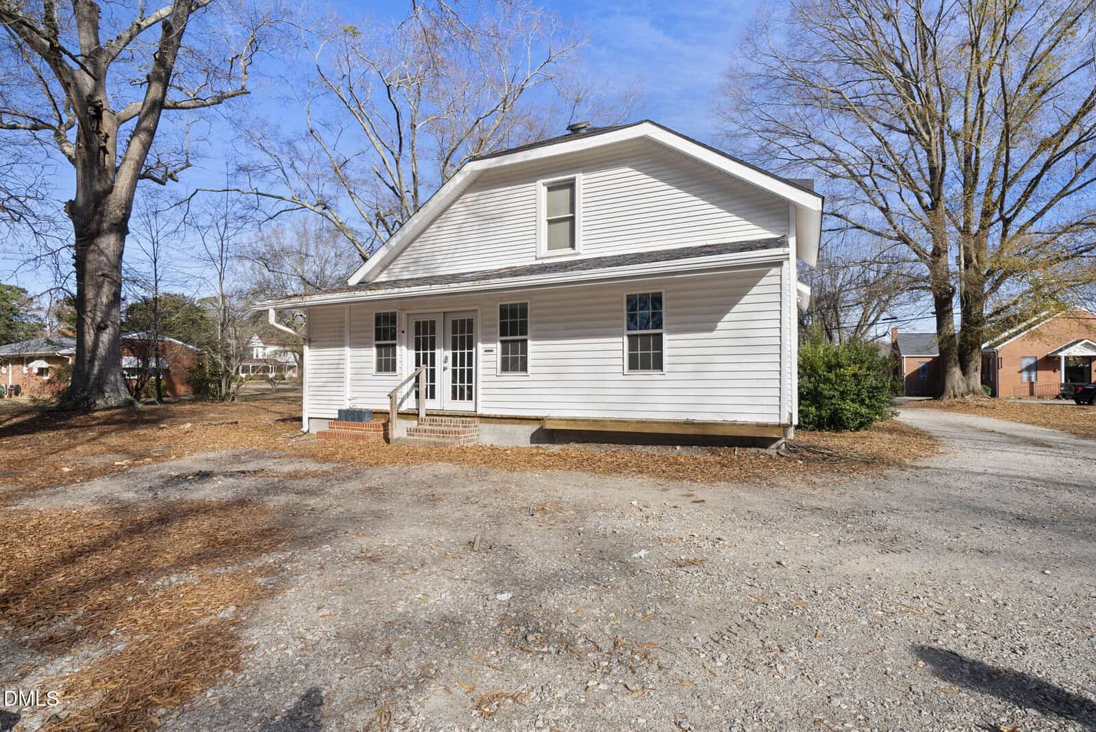 White two-story house with a gambrel roof, multiple windows, and a front entry porch. The yard is mostly dirt and dry leaves, with a few large leafless trees and a clear blue sky in the background.