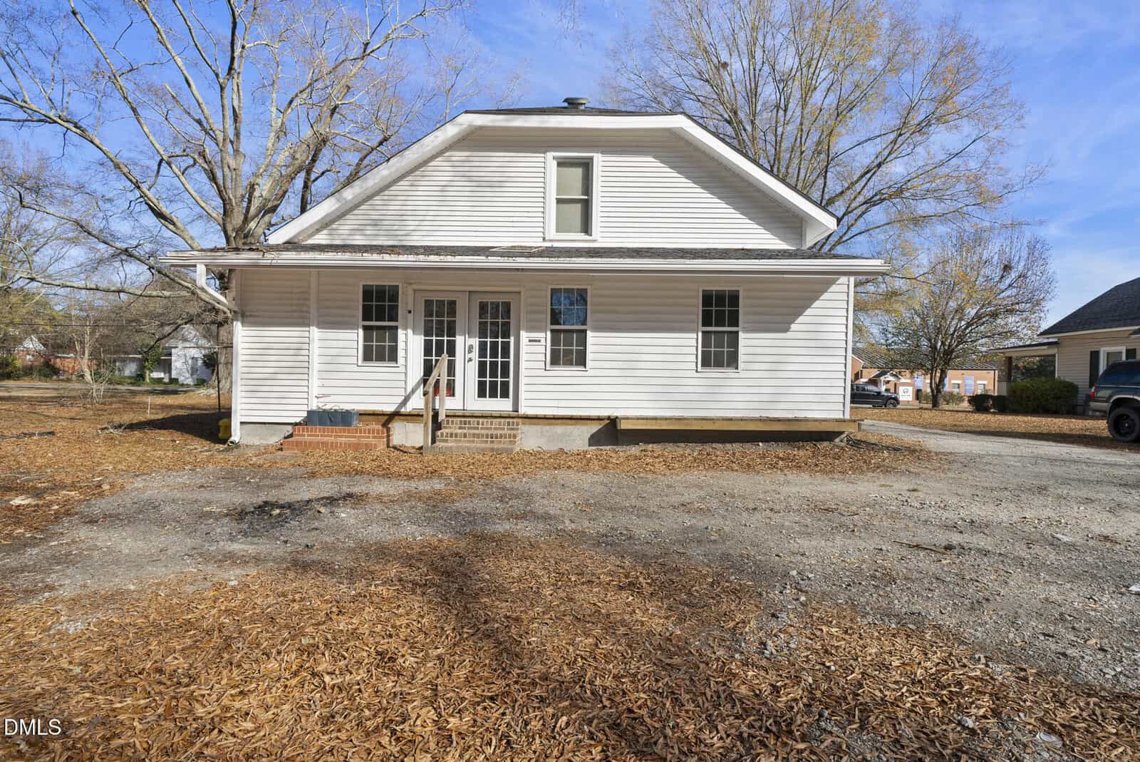 A white, two-story house with a covered front porch, steps leading to the entrance, and large windows. The yard is covered with wood chips and there are bare trees in the background under a clear sky.