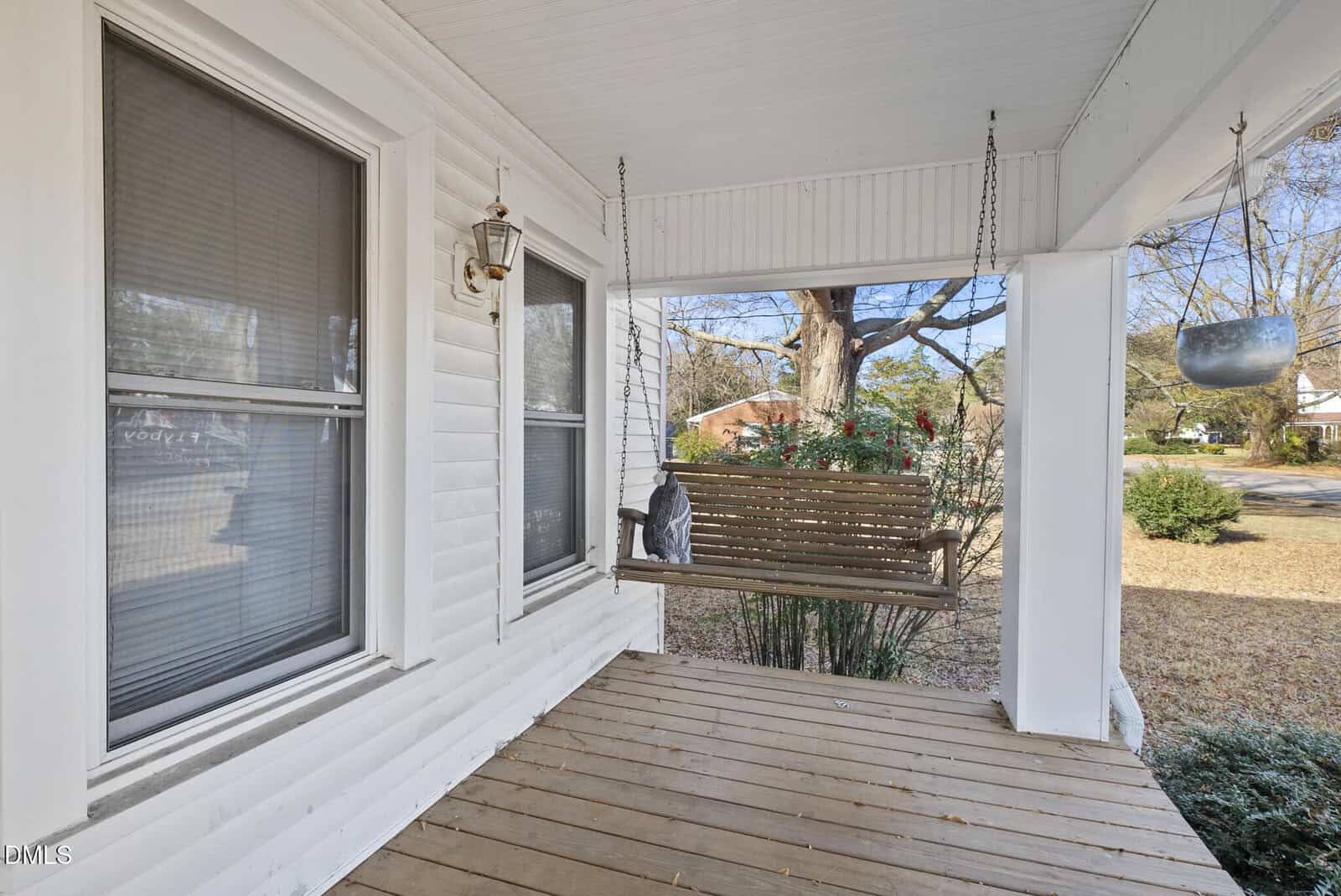 A wooden porch swing hangs on a covered porch with white siding, two large windows, a hanging light, a potted plant, and a view of trees and a residential street in the background.