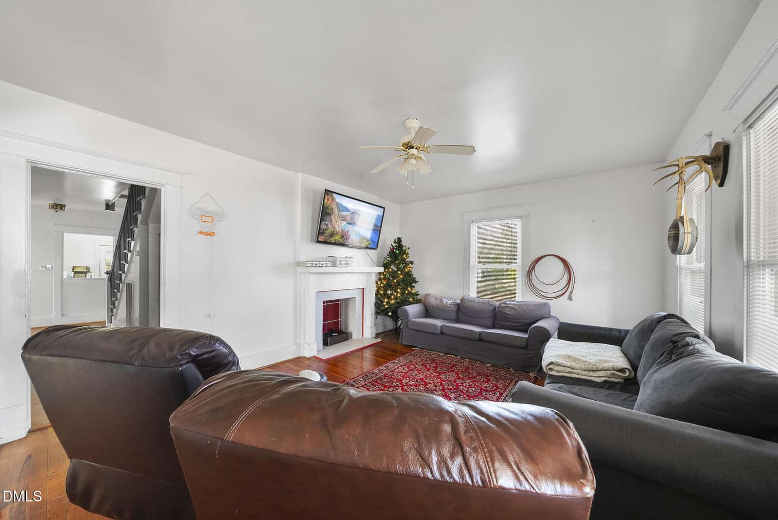 Bright living room with two dark sofas, a leather chair, wall-mounted TV, fireplace, Christmas tree, and large windows. Red rug and ceiling fan add warmth to the white-walled space.