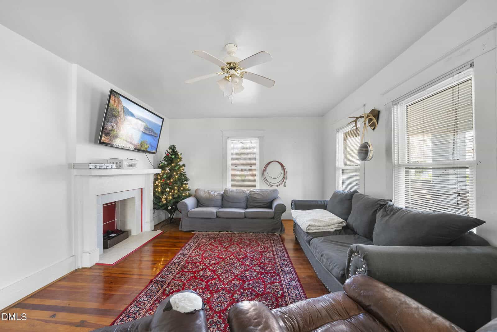 Bright living room with two gray sofas, a red patterned rug, a Christmas tree, wall-mounted TV over a white fireplace, and wooden floors. White walls and several windows let in natural light.