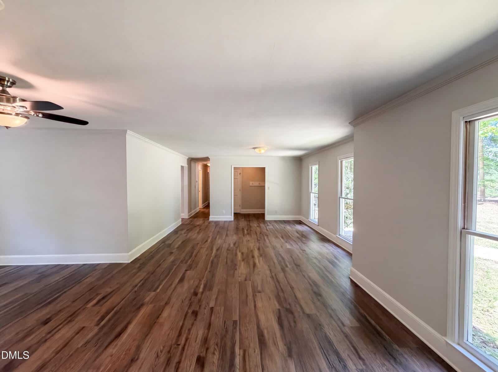 Spacious room with wood flooring, large windows letting in natural light, light gray walls, and a ceiling fan on the left side. A hallway leads to other rooms.