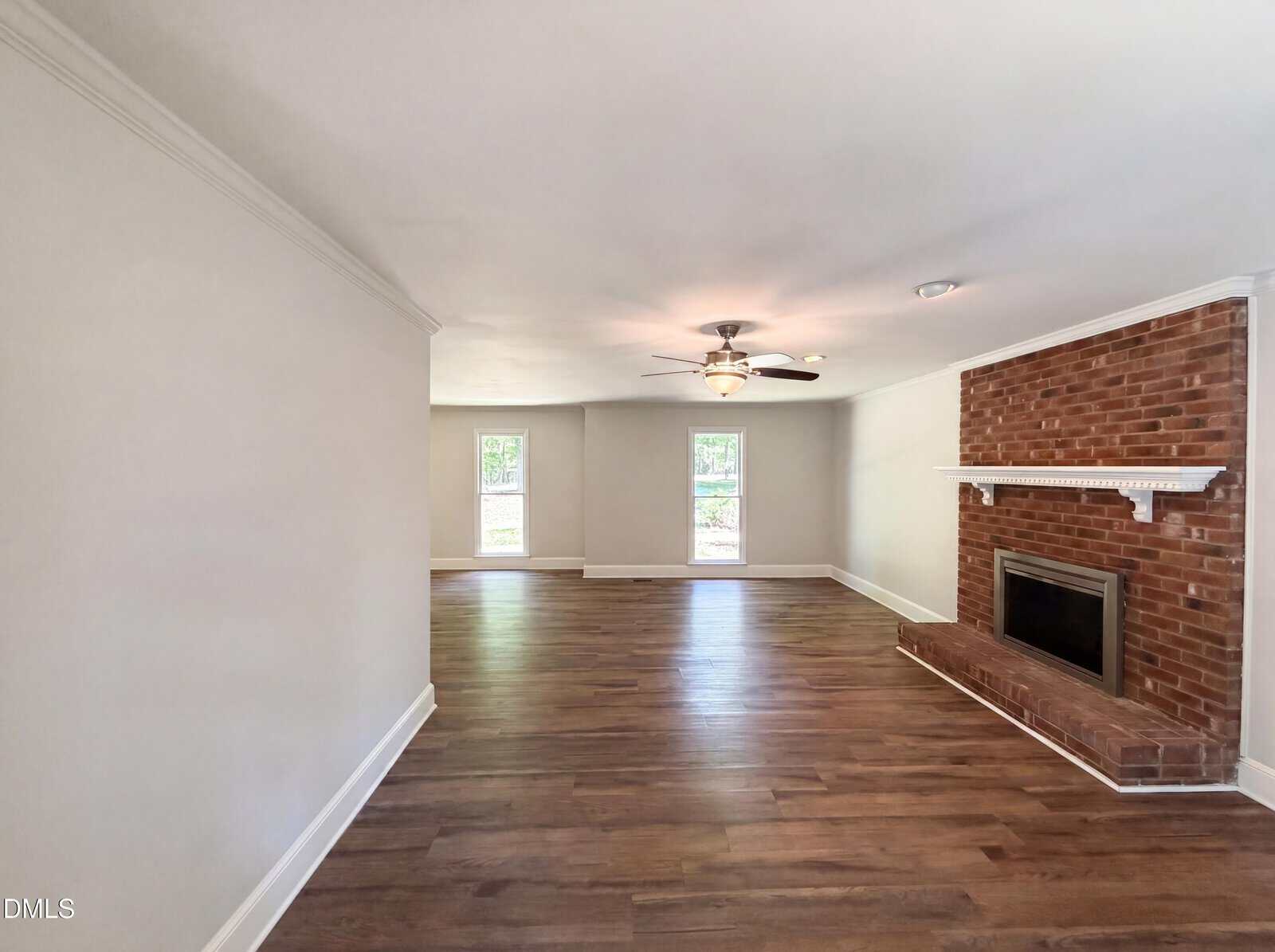 A spacious living room with light walls, wood flooring, a ceiling fan, large windows, and a red brick fireplace with a white mantle.