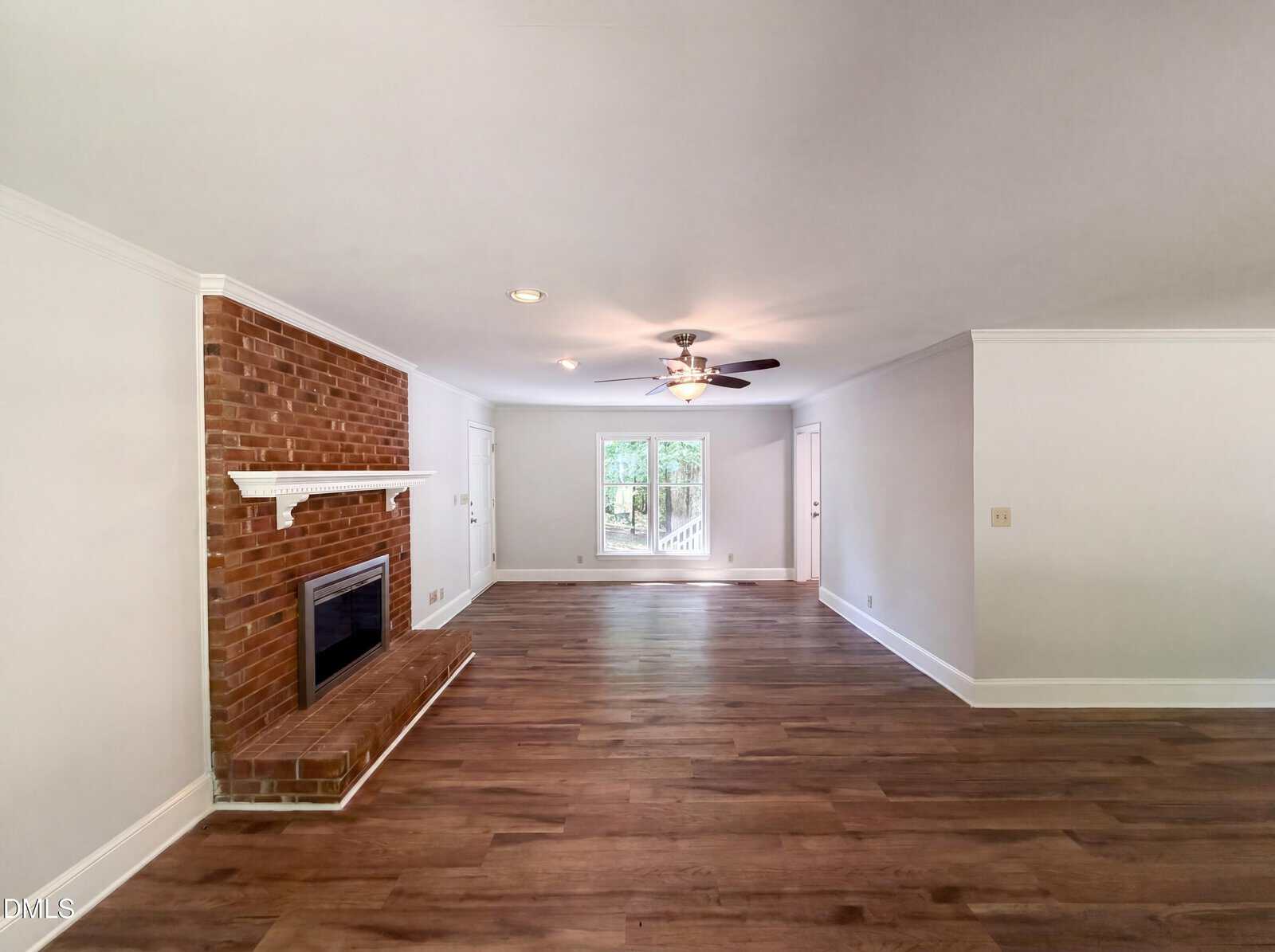 Spacious living room with wood flooring, a brick fireplace on the left, white walls, a ceiling fan with lights, and a large window at the far end letting in natural light.