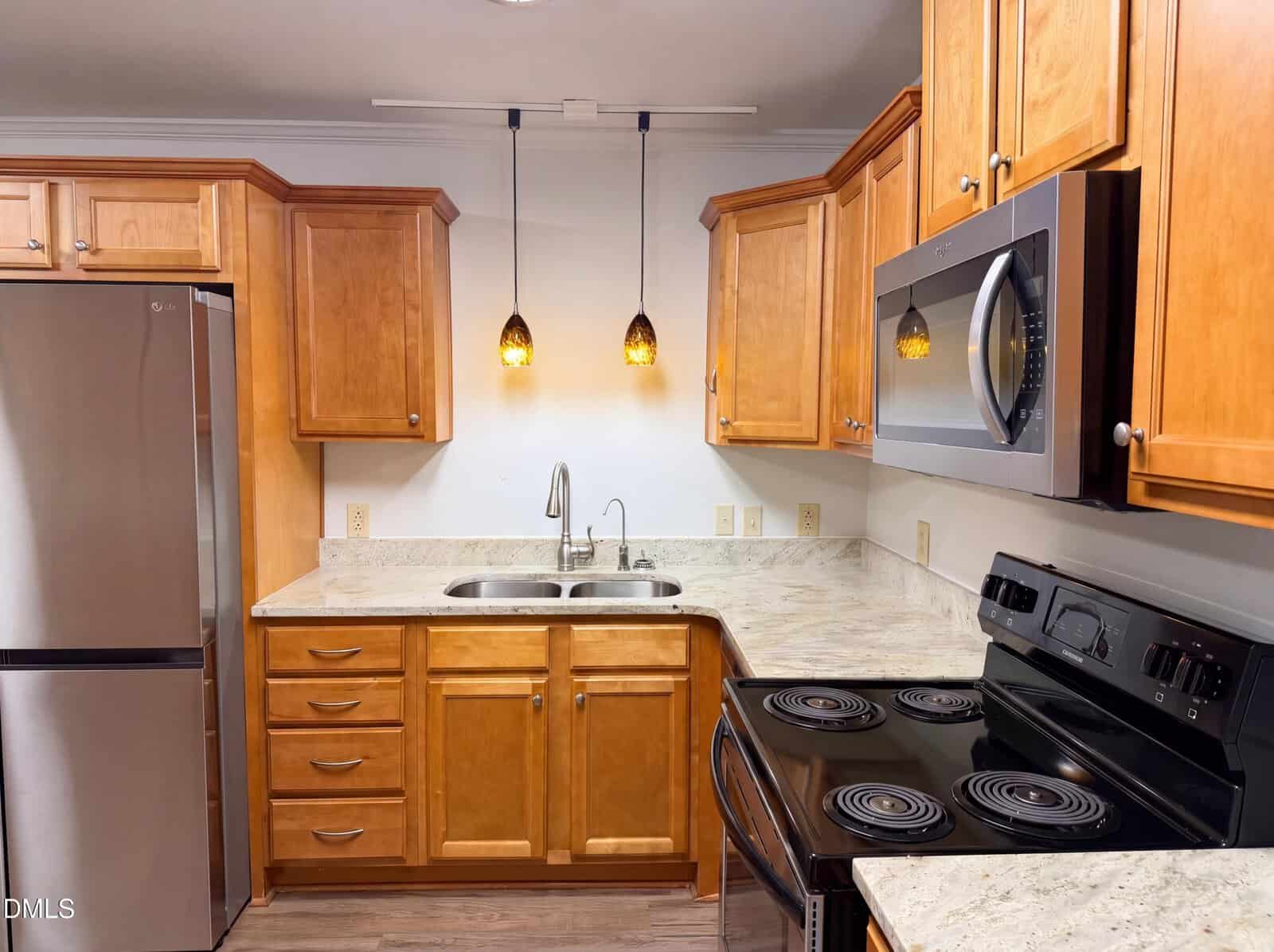 Modern kitchen with light wood cabinets, stainless steel refrigerator, black stove and microwave, double sink, marble countertops, and two hanging pendant lights over the sink area.