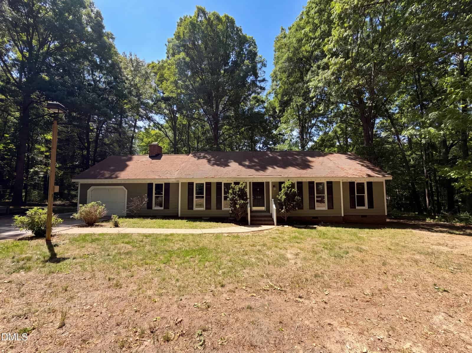 A single-story house with brown siding and a reddish-brown roof sits among tall trees, with a patchy lawn and a driveway leading to an attached garage on the left.