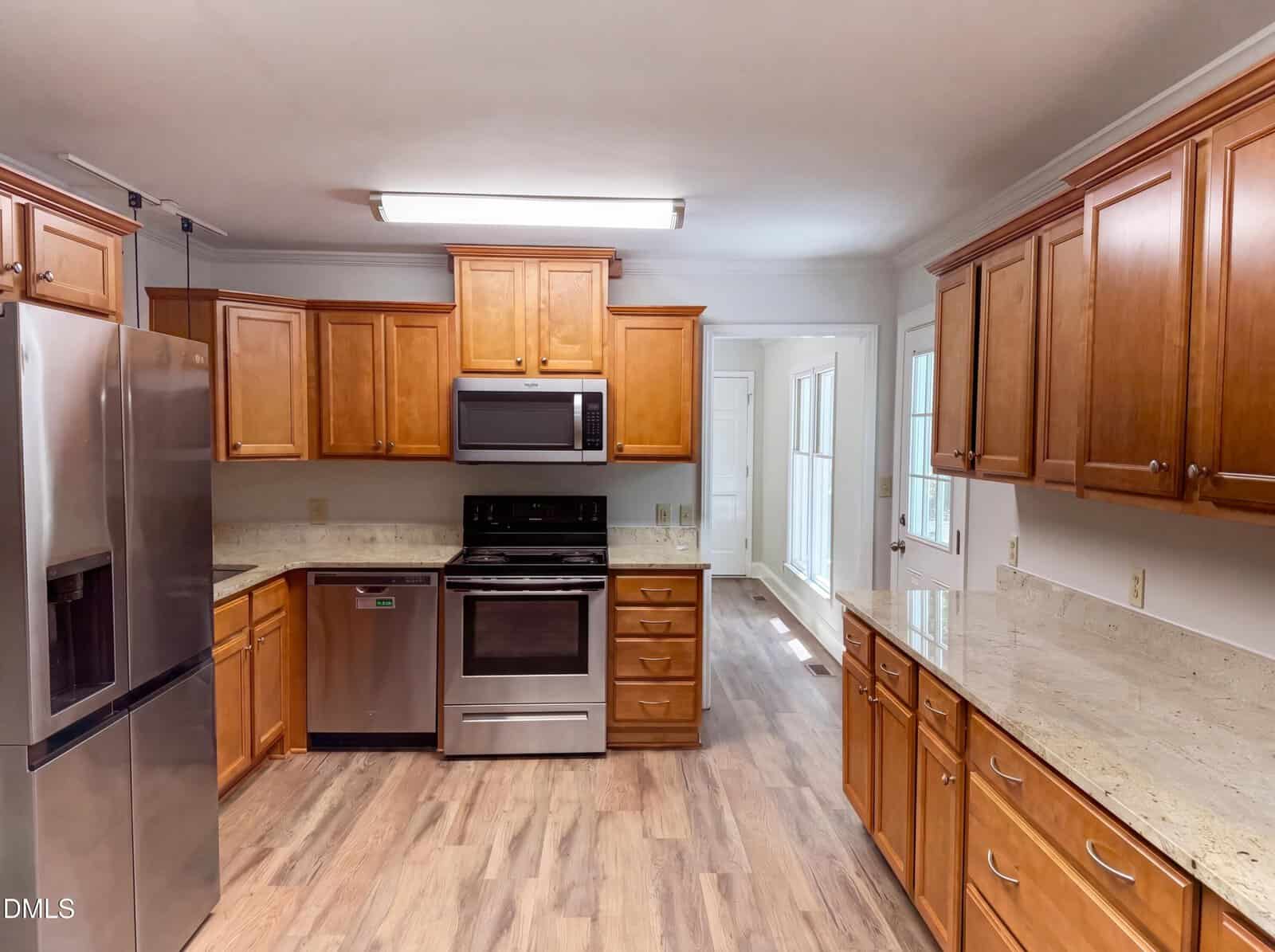 Modern kitchen with wood cabinets, stainless steel appliances, granite countertops, and wood flooring. The room is well-lit with an overhead light and features a bright, open layout with a door and windows in the background.