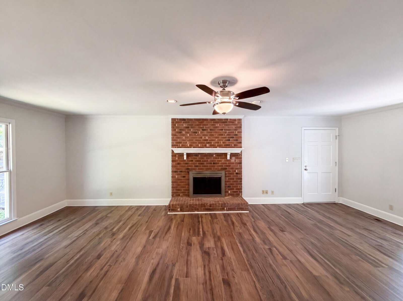 A spacious living room with wood flooring, a ceiling fan, a brick fireplace with a white mantel, light gray walls, and large windows providing natural light. A white door is visible to the right of the fireplace.