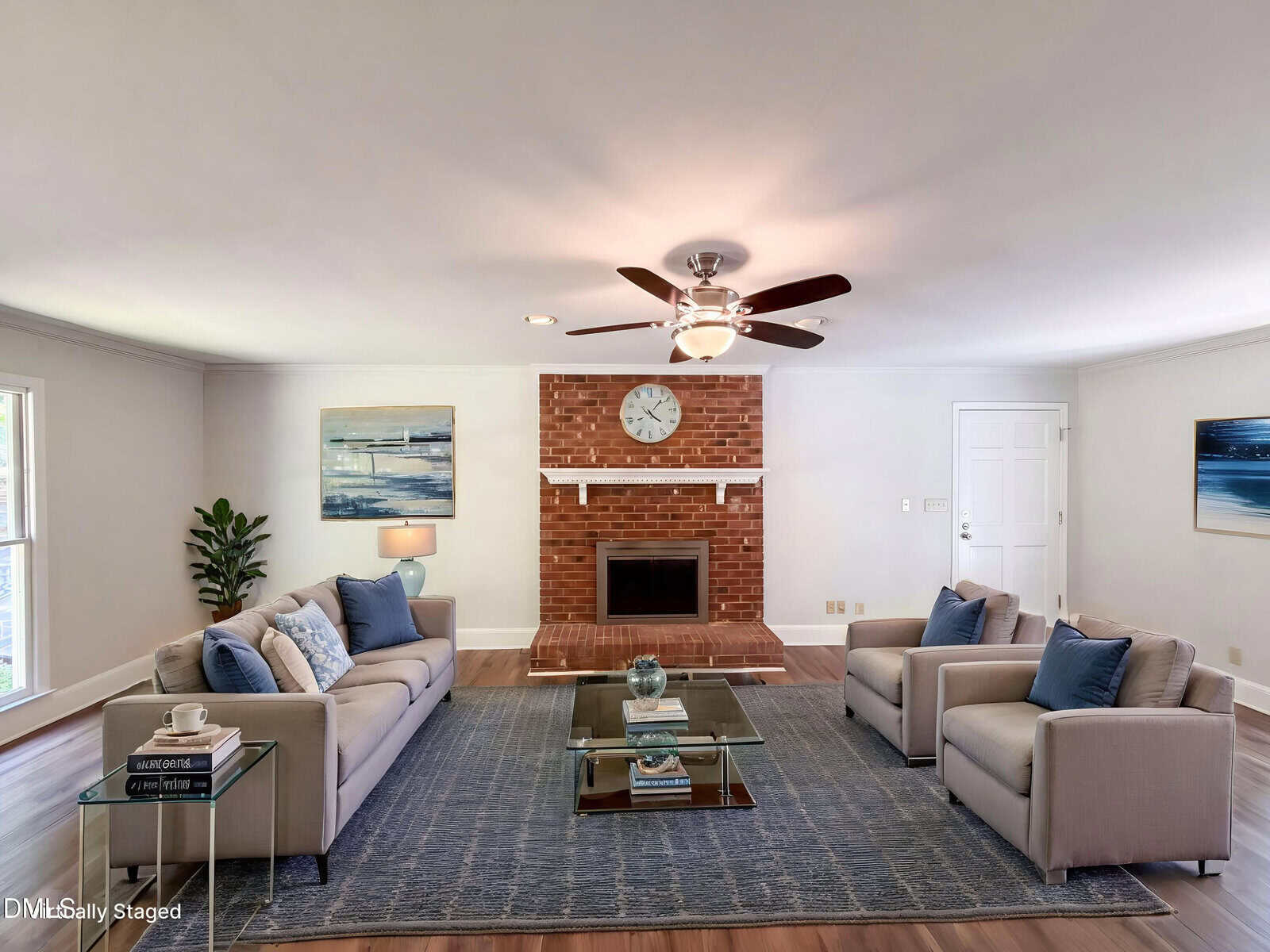A bright living room with a brick fireplace, large clock, and modern decor. Two beige sofas and two chairs face a glass coffee table on a blue rug. Art, plants, and a ceiling fan complete the cozy, staged space.