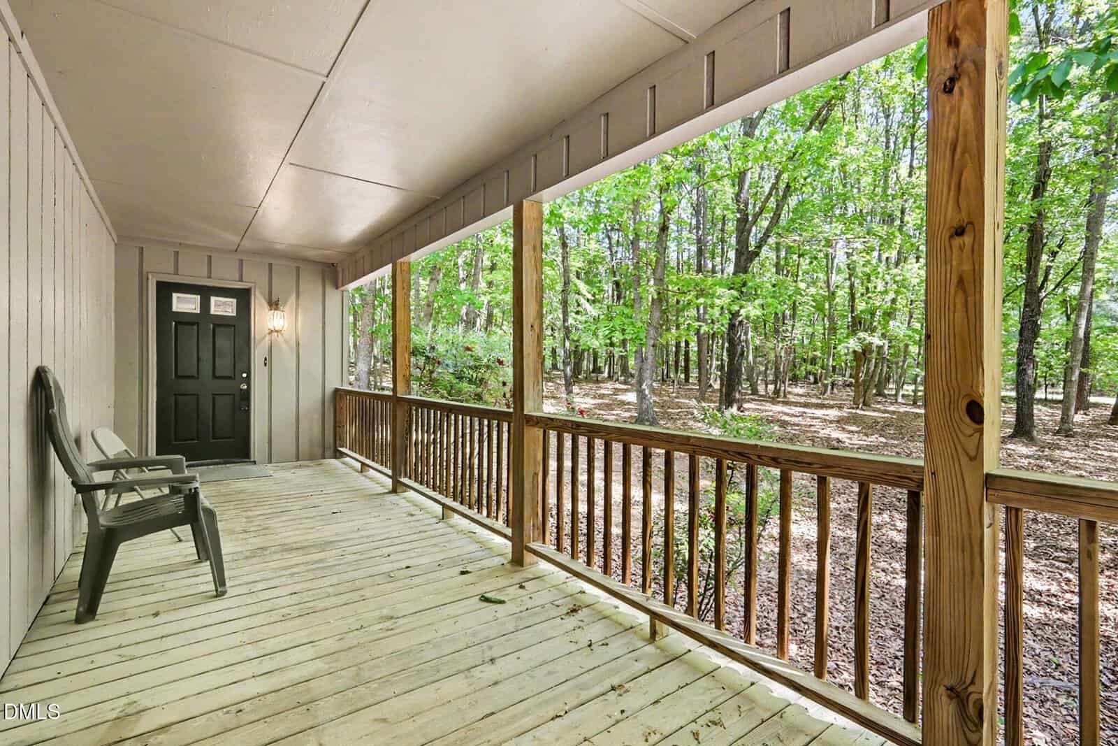 A spacious wooden front porch with railings, two chairs, and a wall-mounted light overlooks a forested yard with tall trees and scattered leaf litter. The porch leads to a black front door.