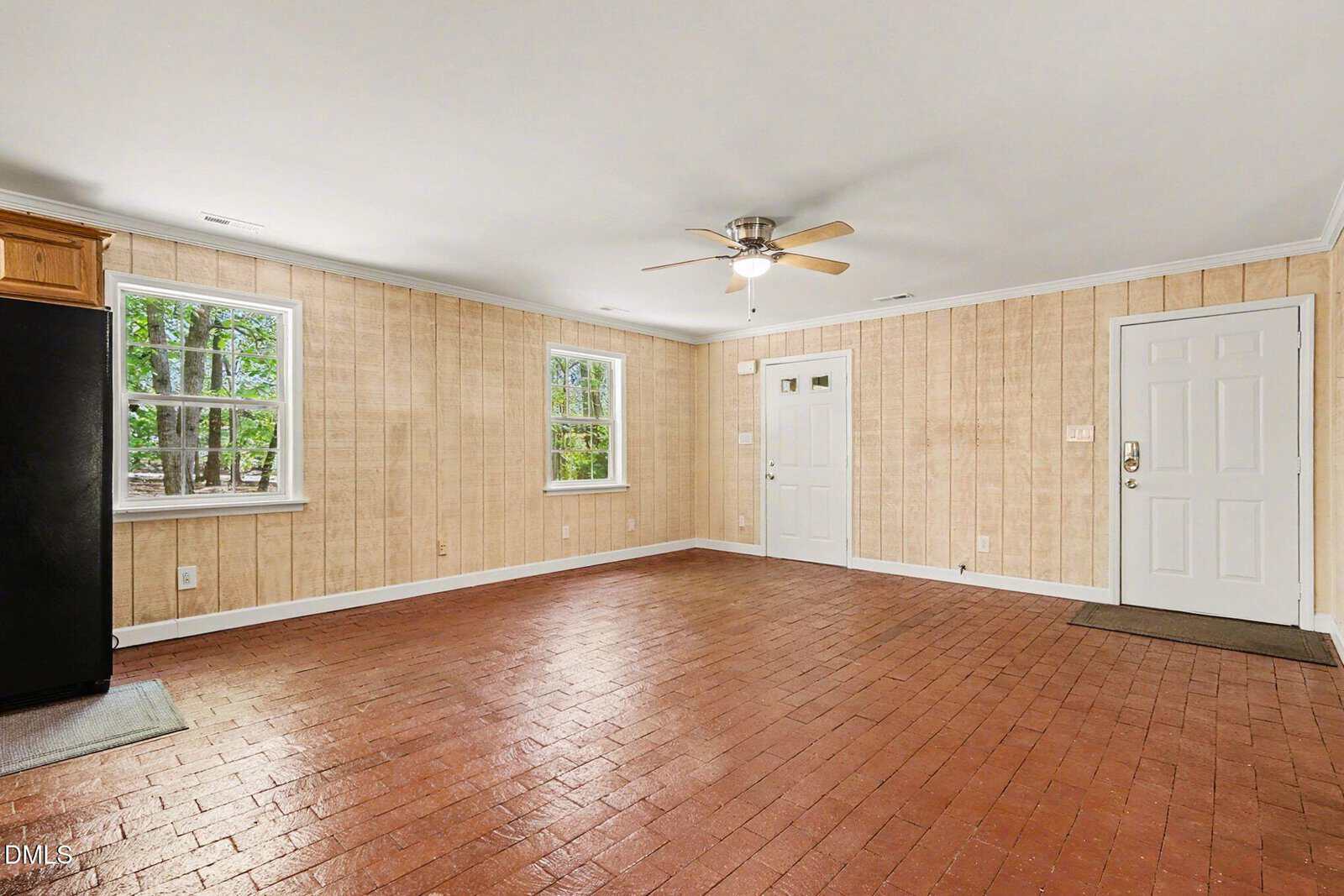 A spacious room with brick flooring, wood-paneled walls, two windows, a ceiling fan with light, a white door, and a black refrigerator in the corner.