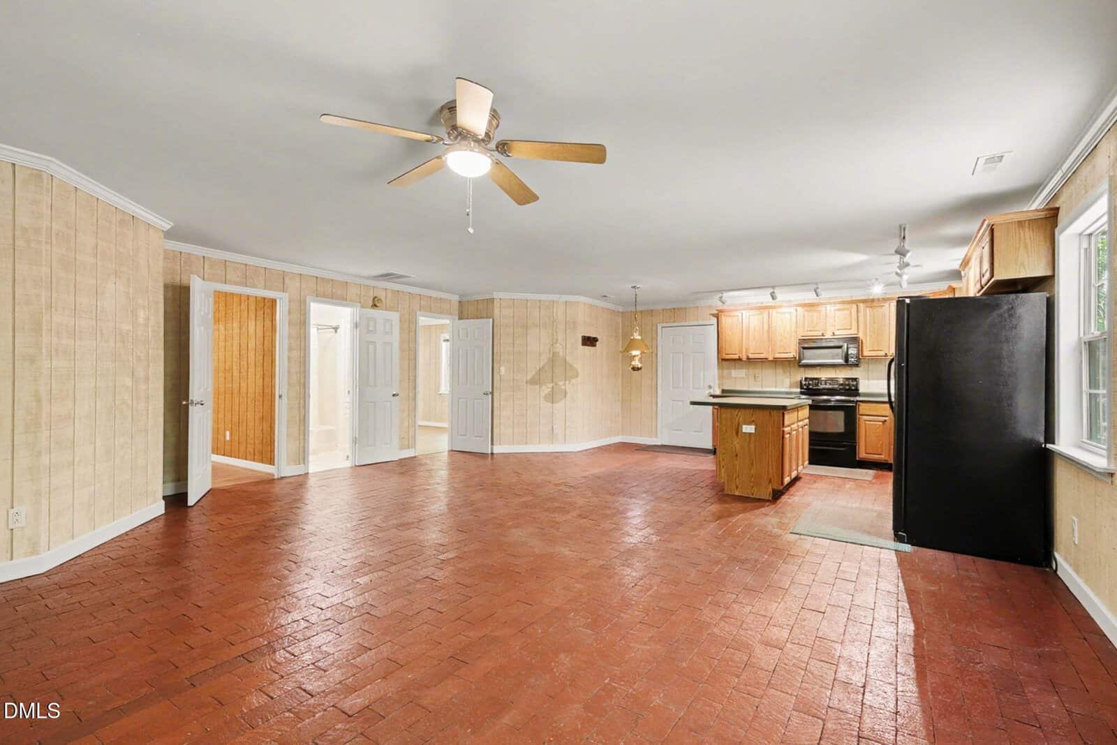 Spacious open-plan room with red brick tile flooring, light wood-paneled walls, ceiling fan, and a kitchen area featuring wooden cabinets, black appliances, and island; multiple doors lead to other rooms.