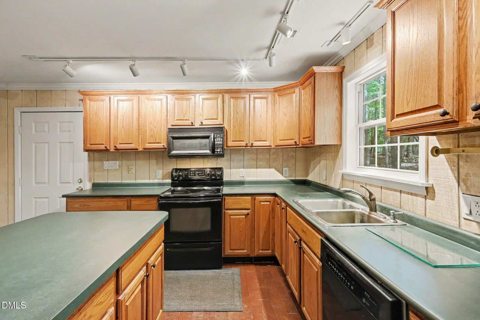 A kitchen with wood cabinets, green countertops, black appliances including a stove, microwave, and dishwasher, a double sink under a window, and track lighting overhead.