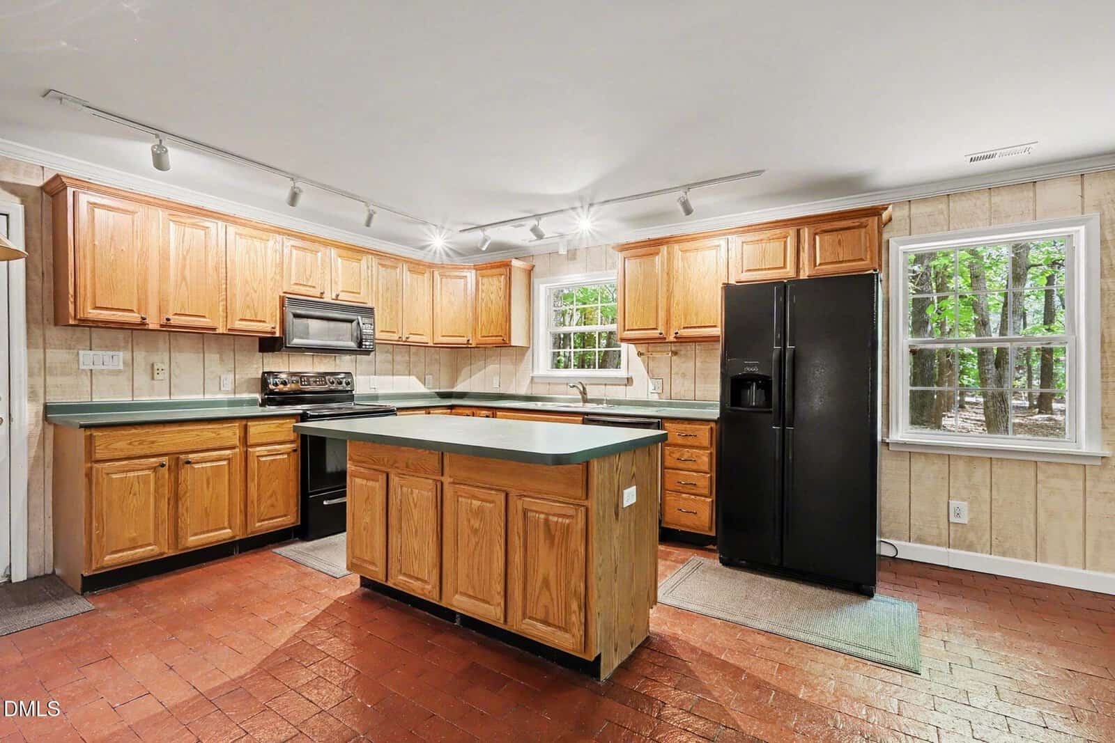 Spacious kitchen with wooden cabinets, black appliances, green countertops, a center island, brick floor, large windows, and track lighting.