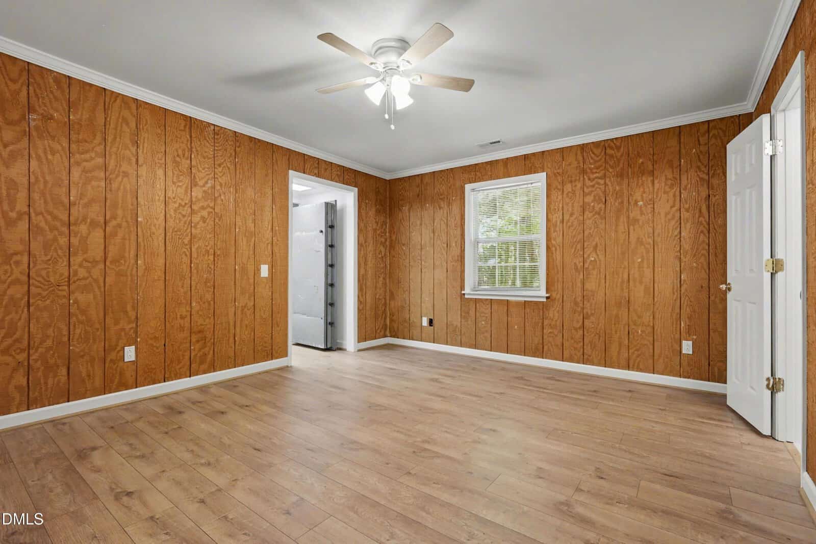 Empty room with wood-paneled walls, wood flooring, a ceiling fan with lights, a window with blinds, white trim, and an open doorway leading to another room.