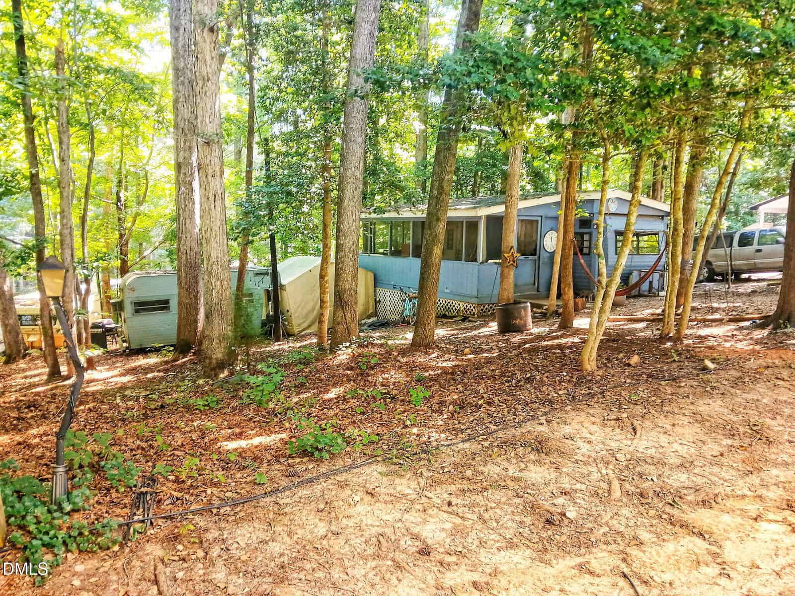 A blue mobile home and two white trailers are situated among tall trees in a wooded area with sunlight filtering through the leaves. A dirt ground covers the foreground and some cars are partially visible in the background.