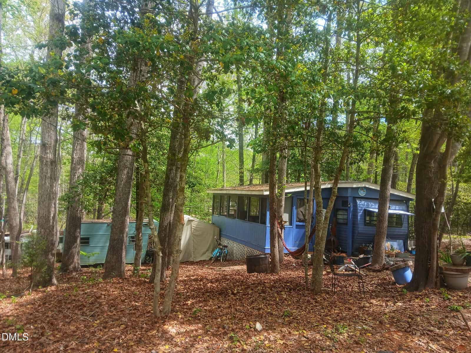A small blue cabin with a screened porch is nestled among tall trees in a wooded area, surrounded by fallen leaves and various outdoor items, including a covered object and a blue trailer.