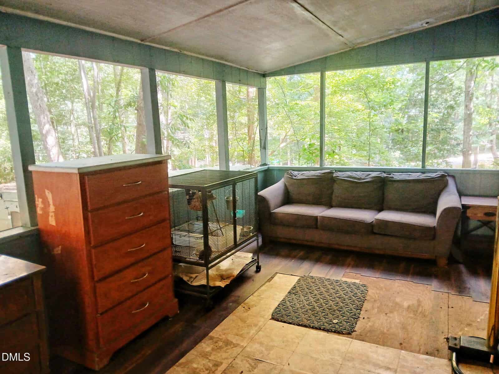 A cozy sunroom with large windows showing trees, featuring a gray couch, a wooden dresser, a small rug, and a birdcage on dark wood flooring. Natural light fills the space.