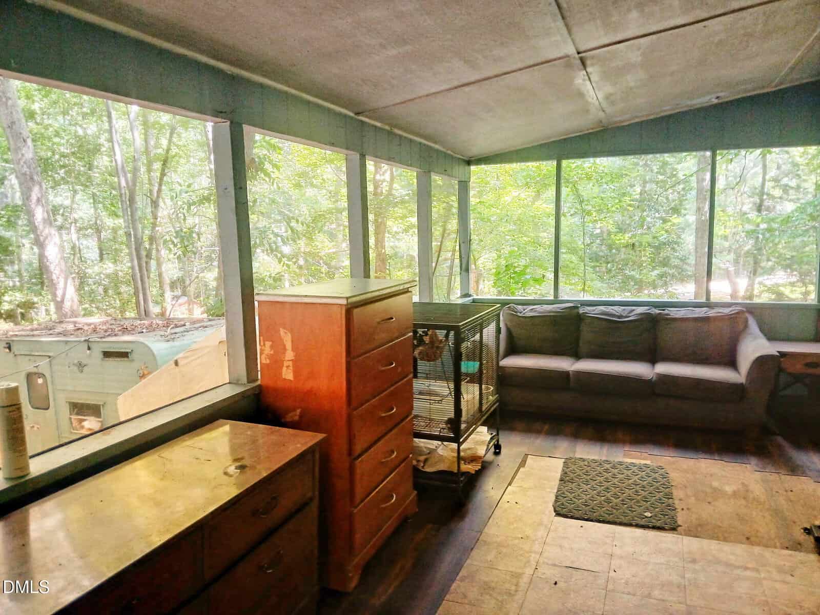 A sunroom with large windows, a gray sectional sofa, wooden dressers, a birdcage, and a view of trees outside. The floor has a rug and some worn spots. Light fills the room.