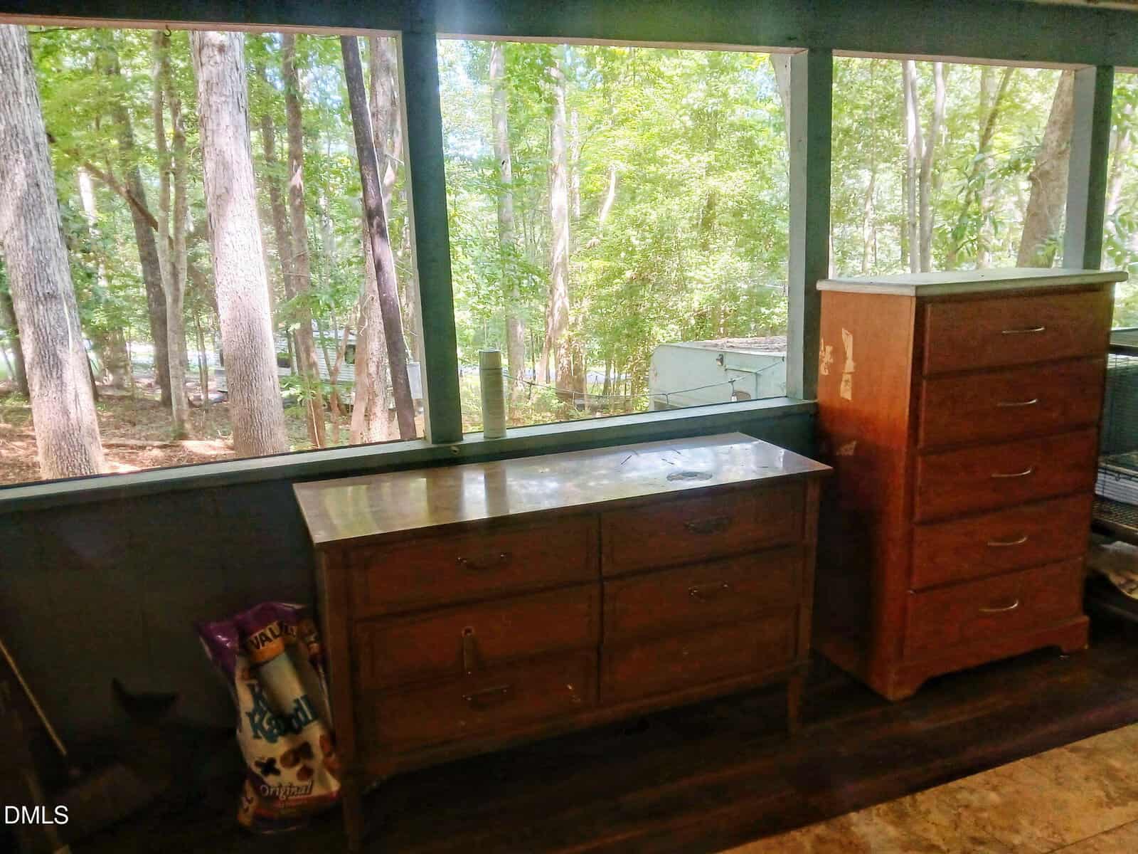 Two wooden dressers sit on a covered porch with screened windows. A bag of cat litter is on the floor next to the dressers. Trees and greenery are visible outside through the screens.