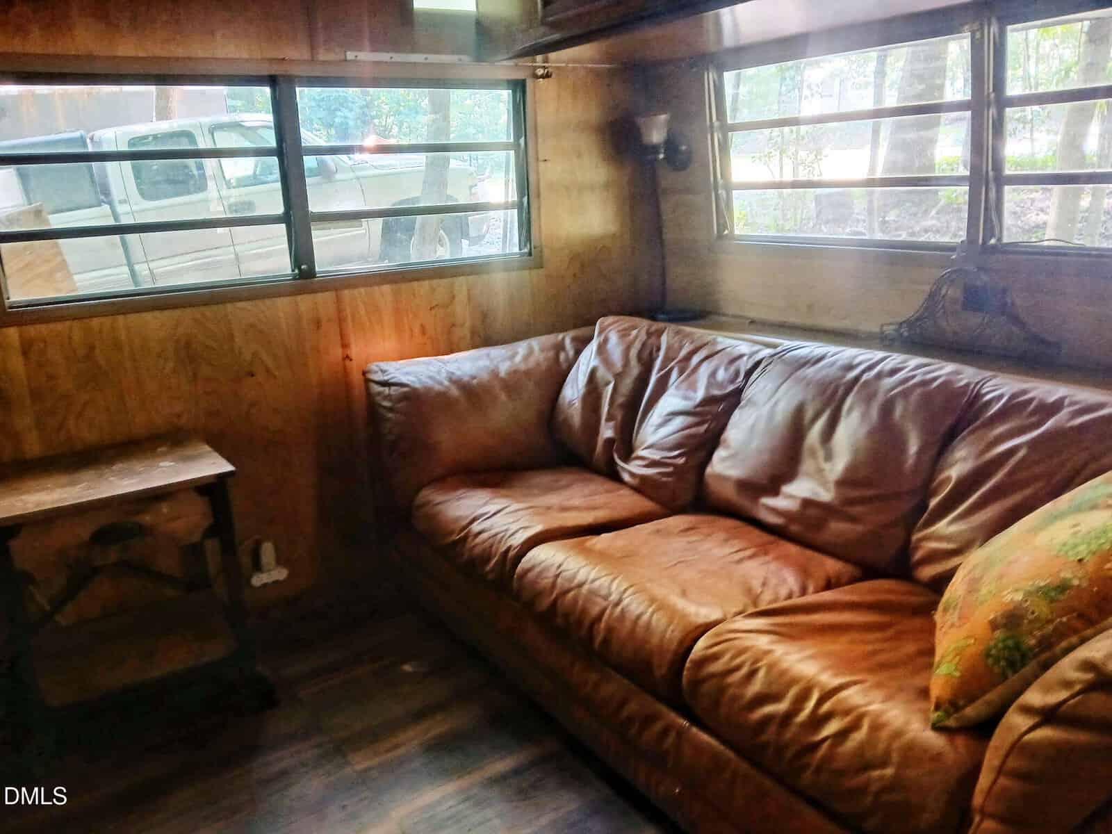 A brown leather couch with a patterned pillow sits in a wood-paneled room with large windows. Sunlight streams in, and a truck is visible outside through the windows. A small wooden side table is nearby.