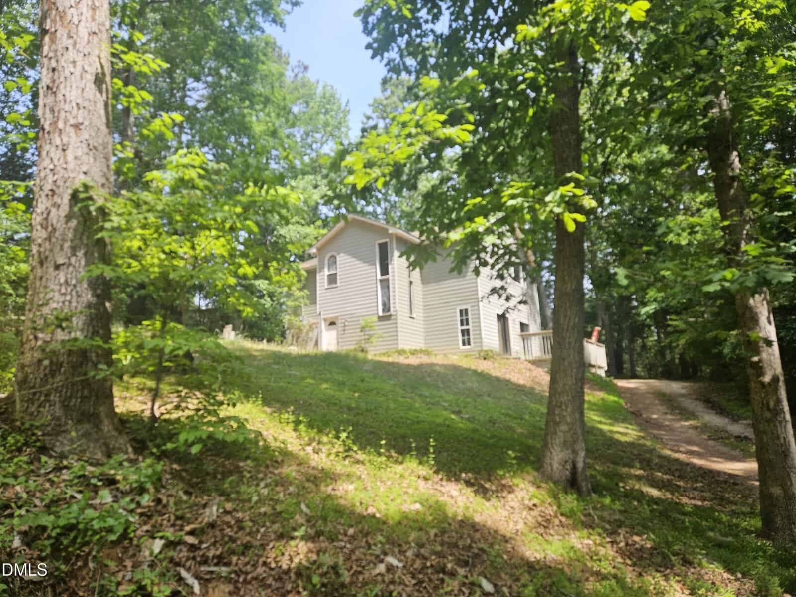 A light gray house with white trim sits on a grassy hill surrounded by tall trees and greenery, with a dirt driveway leading up to the home. Sunlight filters through the leaves, creating a peaceful, wooded setting.