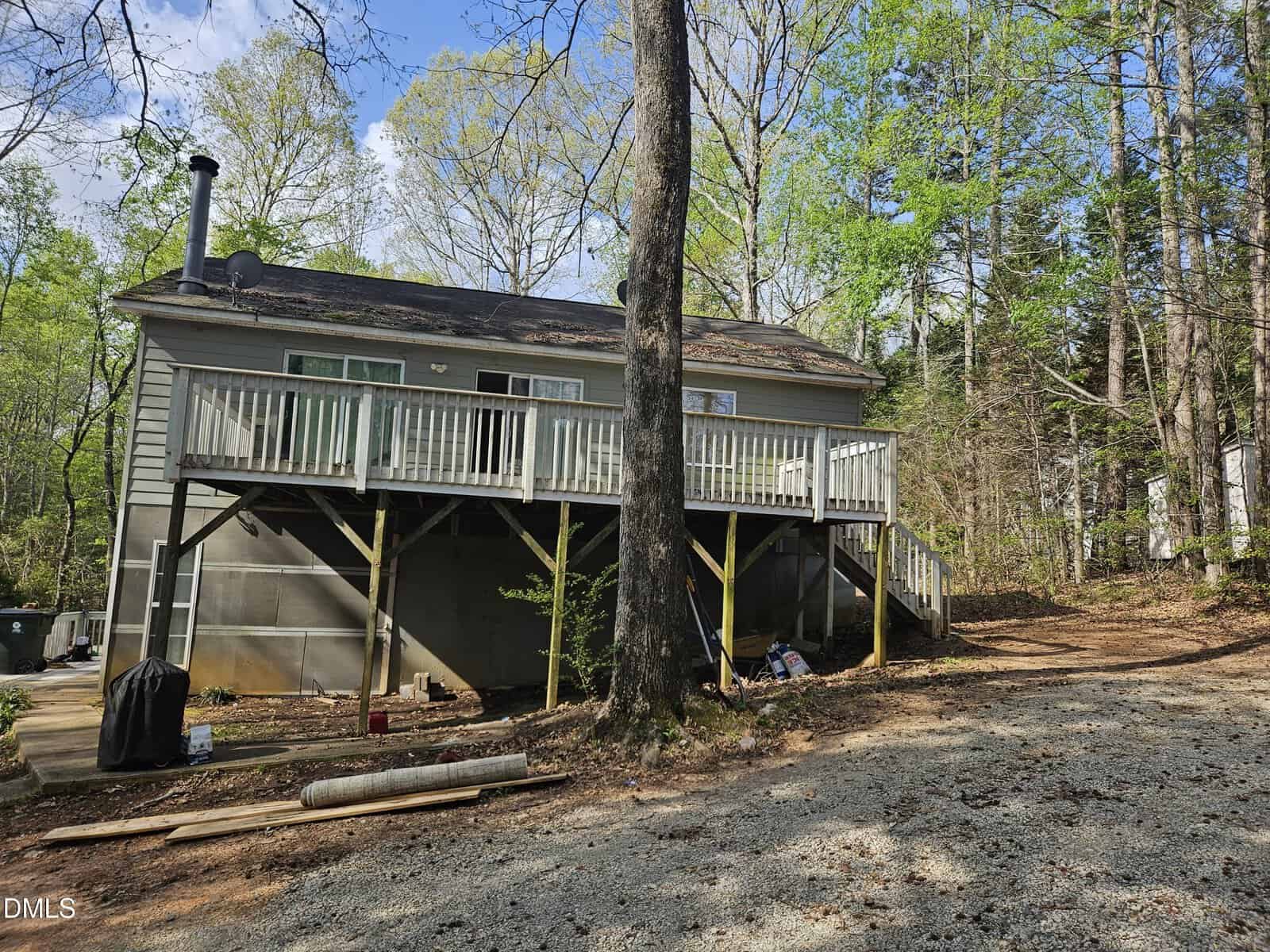 A small, elevated house with a wooden deck and stairs sits among tall trees. The ground below is gravel and dirt, with some construction materials scattered nearby. The setting is wooded and peaceful.